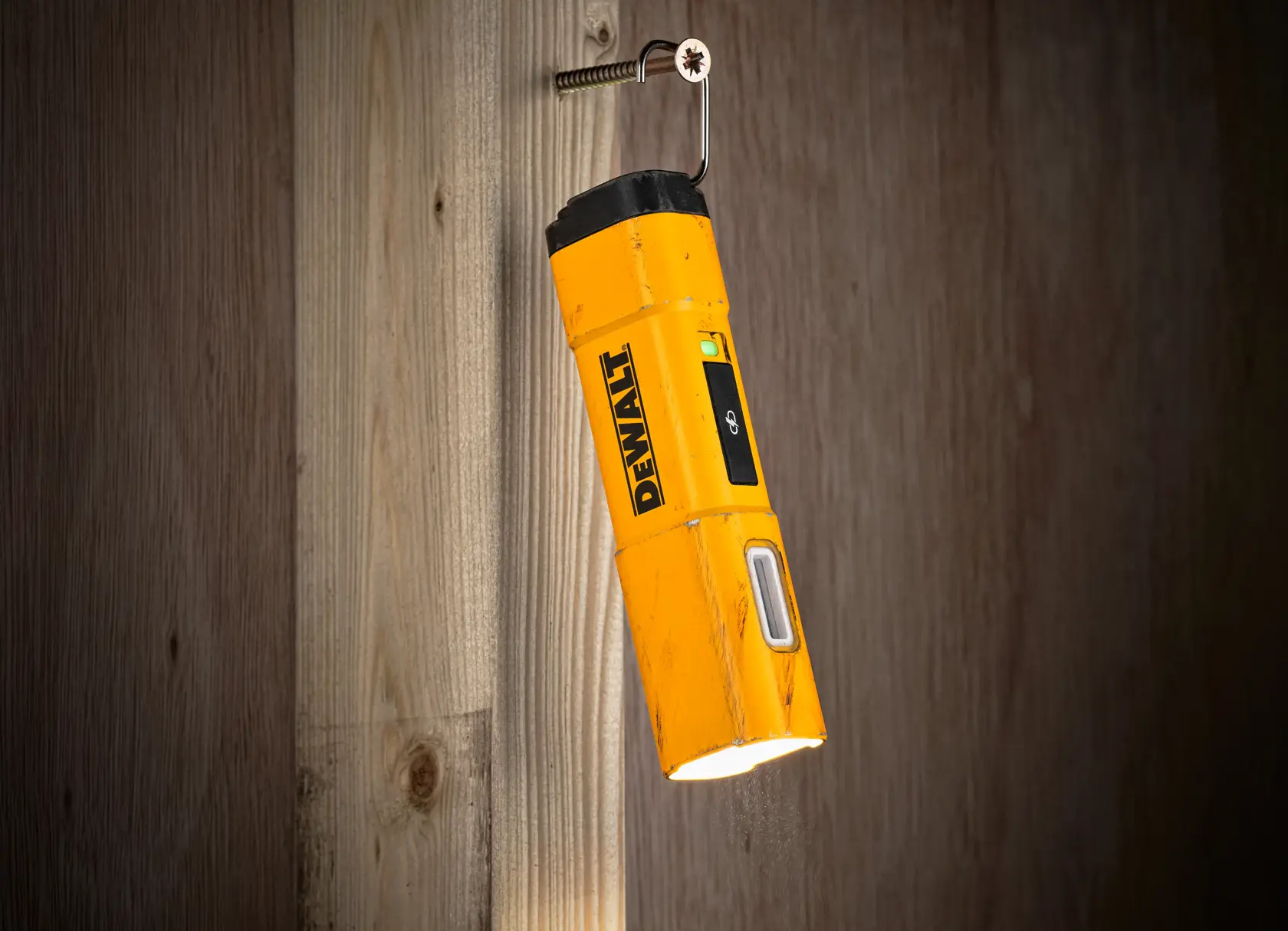 A yellow DEWALT DCL183 portable LED flashlight is hanging from a hook screwed into a wooden wall, illuminating downward.