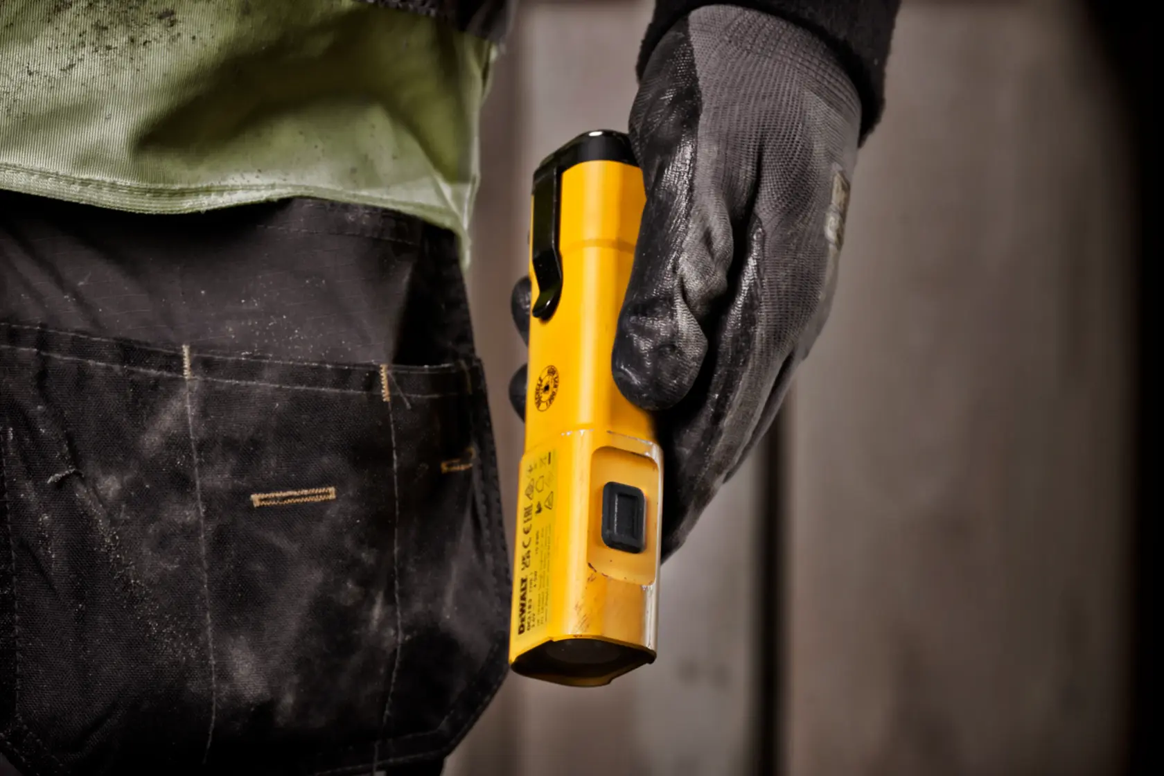 A person wearing black work gloves and work pants holds a yellow DEWALT DCL183 flashlight at their side.