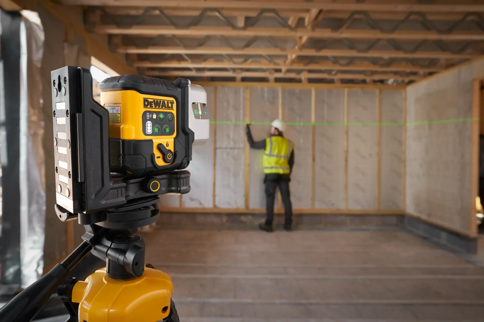 DEWALT laser level on tripod projects green line in construction site, worker marks wall alignment.