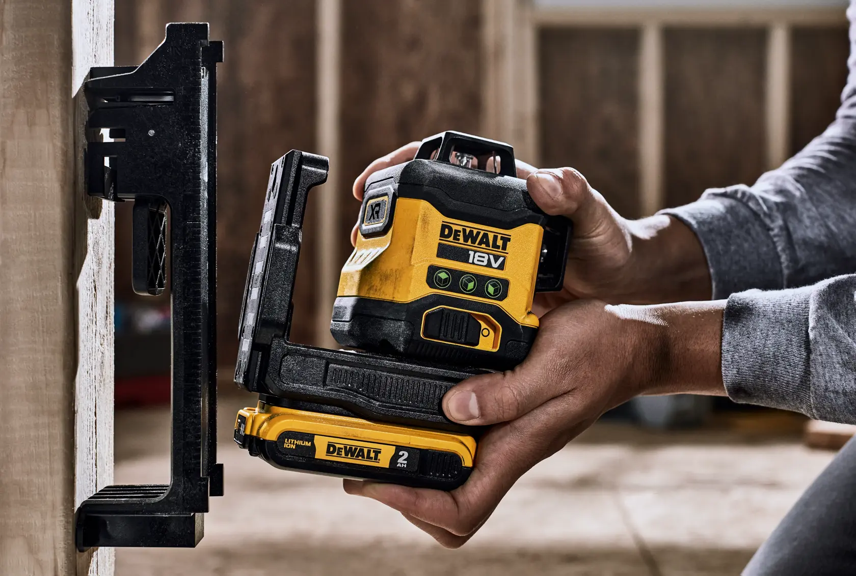 Primer plano de manos colocando un nivel láser DEWALT 18V, modelo DCLE34031N, contra una superficie vertical en el interior de una obra de construcción. El dispositivo es amarillo y negro con etiquetas visibles y compartimento de batería a la vista.