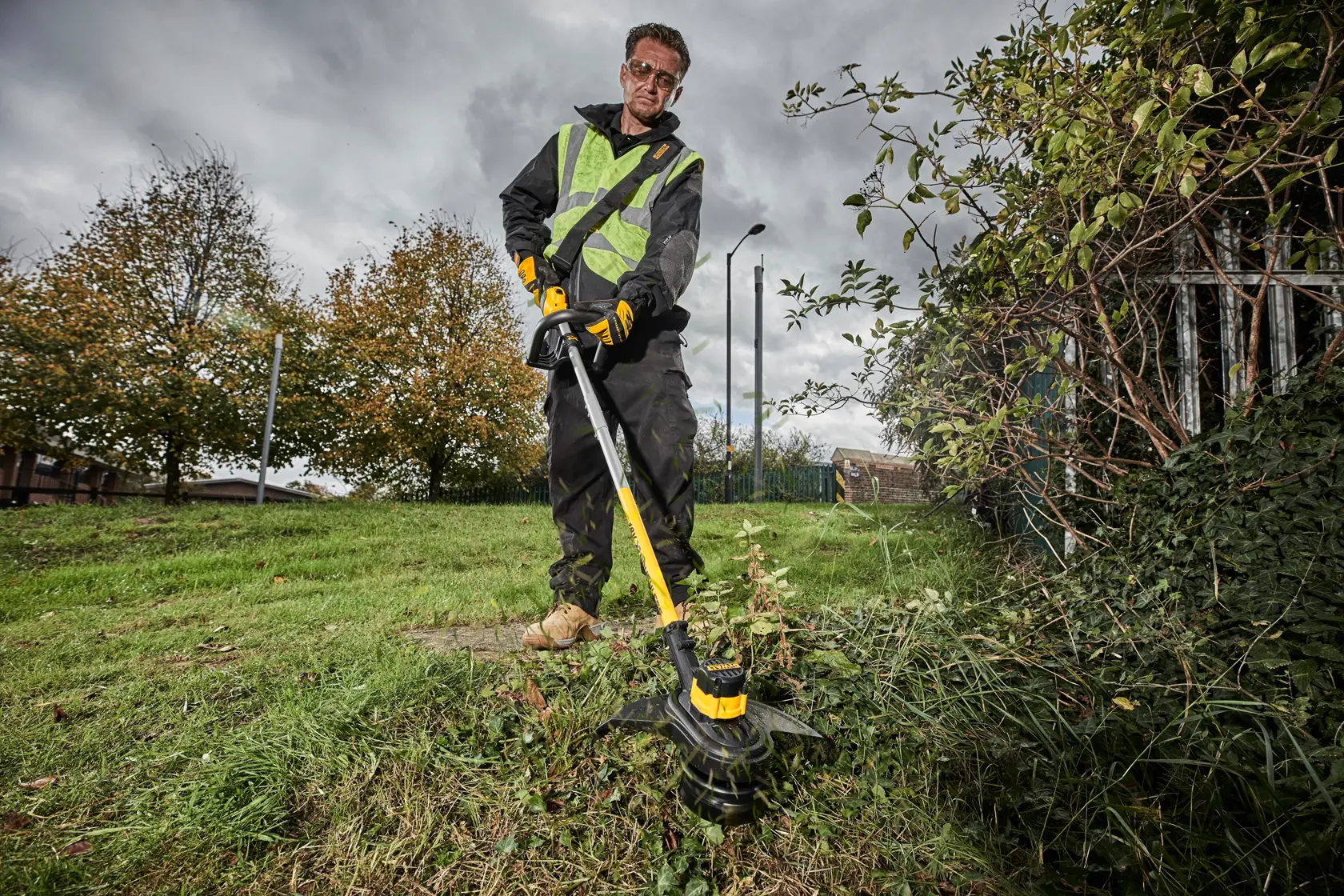 Person using a DEWALT grass trimmer outdoors, wearing work gloves and a safety vest.