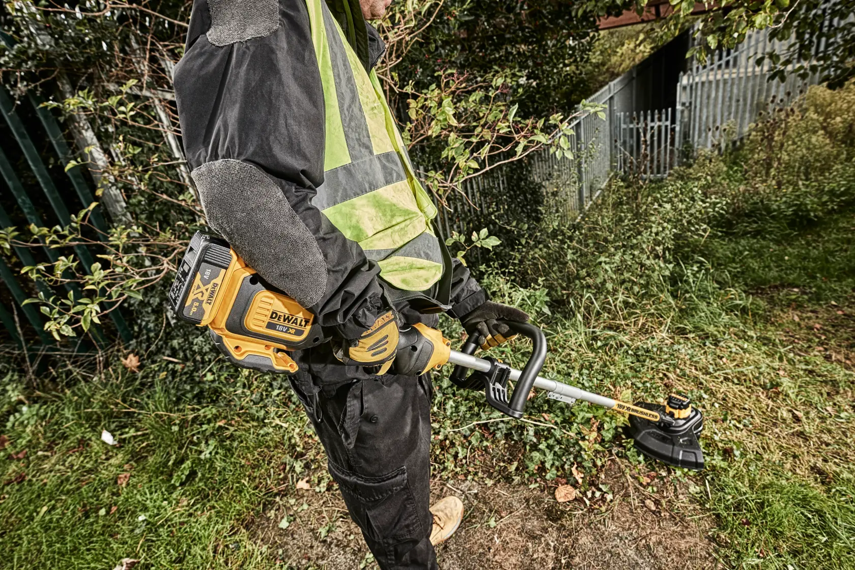 Person using a DEWALT DCM561PBS string trimmer outdoors, cutting grass near a fence and bushes.