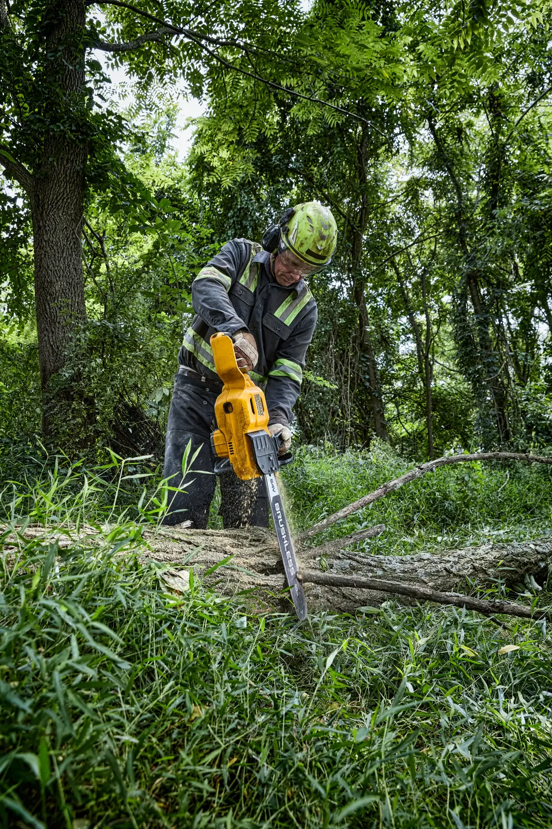 A person wearing protective gear uses a yellow DEWALT cordless chainsaw to cut a fallen tree in a forested area.
