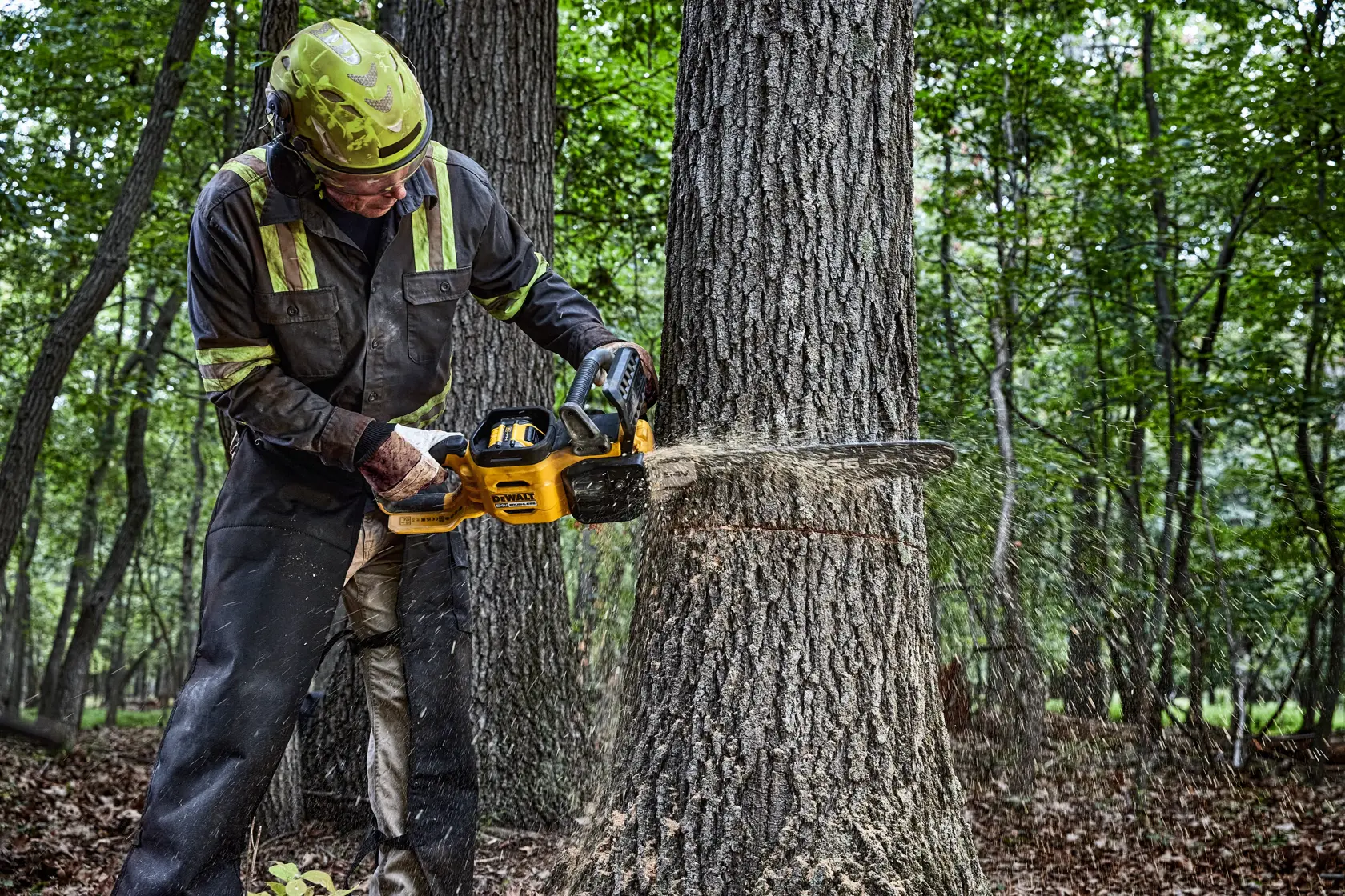 A person wearing protective gear operates a yellow DEWALT chainsaw while cutting into a tree trunk in a forest setting. Sawdust is visible as the chainsaw cuts through the wood.