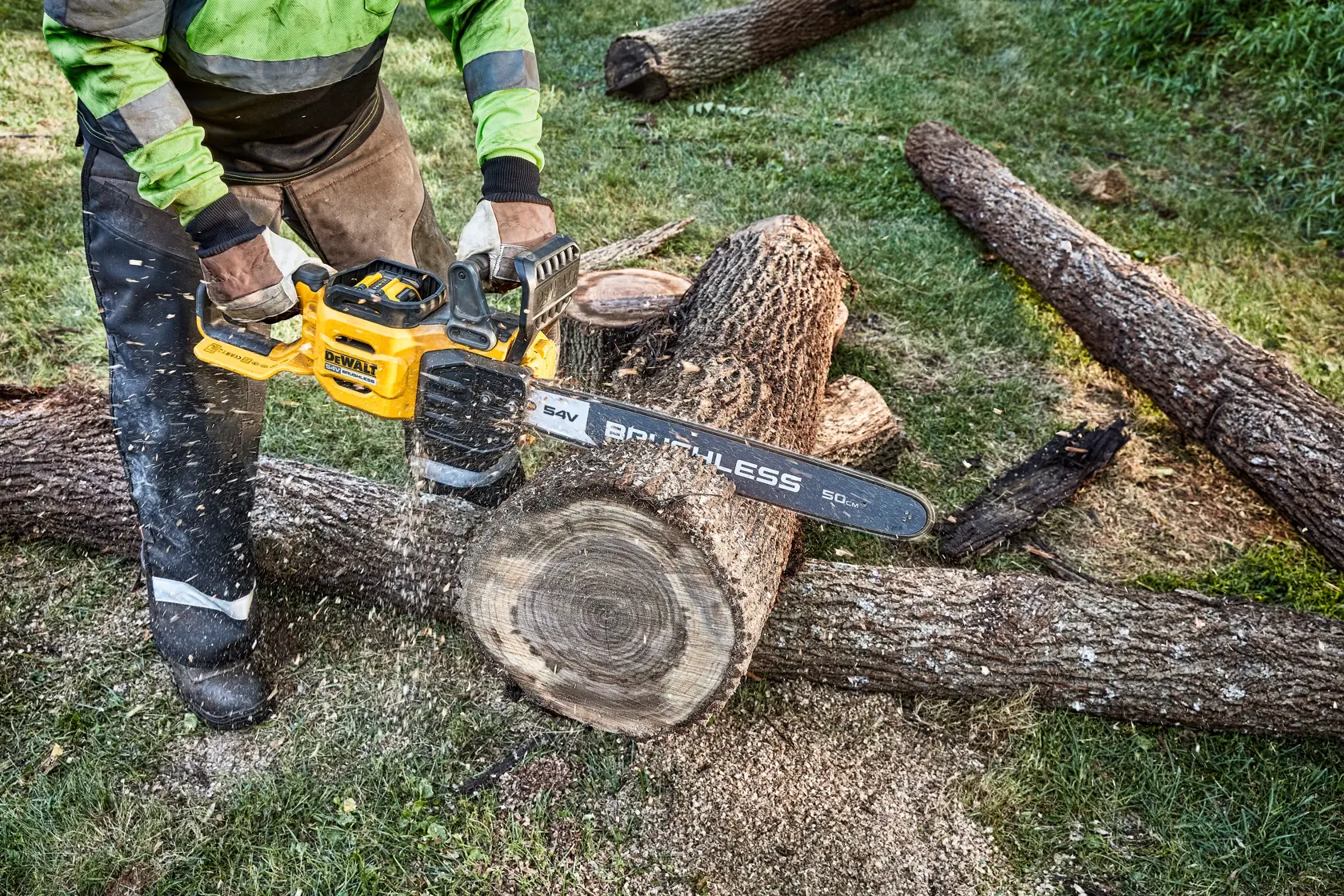 A person using a yellow DEWALT cordless chainsaw to cut through a large log outdoors. The chainsaw is in the process of cutting through the wood, producing wood shavings.