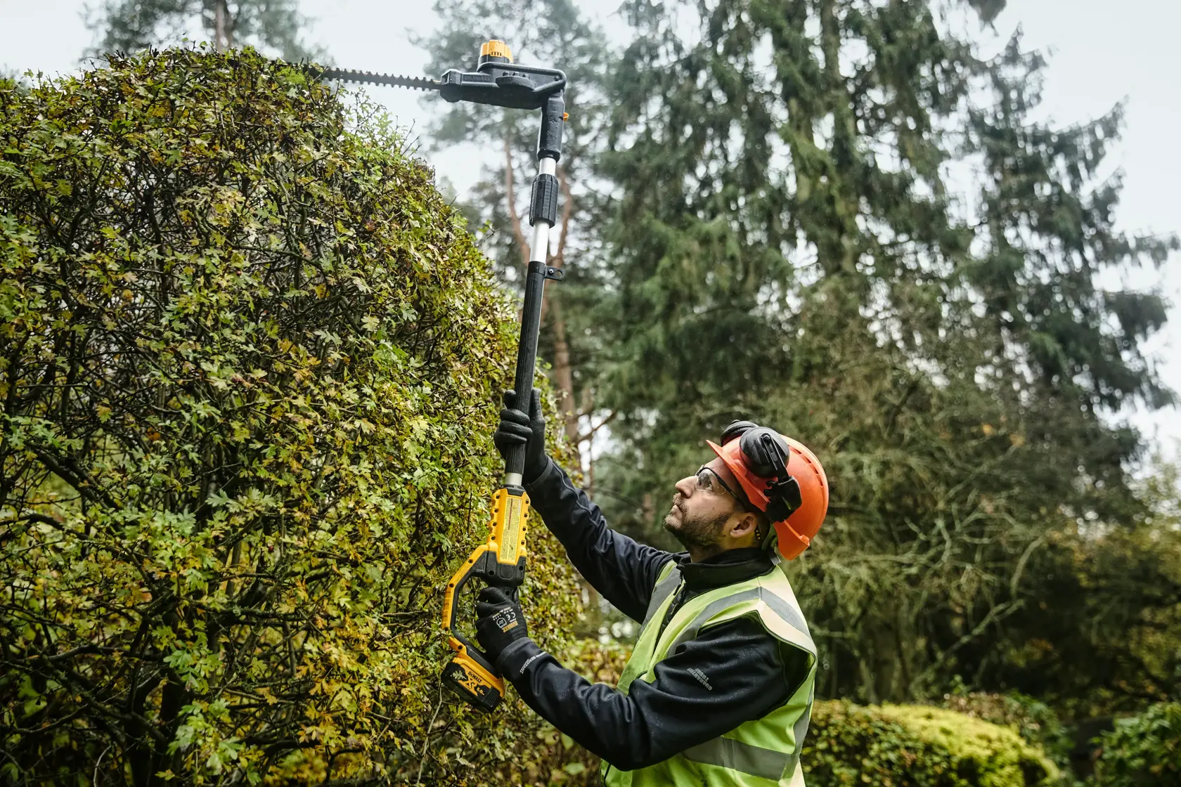 Persona usa cortasetos telescópico DEWALT inalámbrico al aire libre y lleva equipo de seguridad.