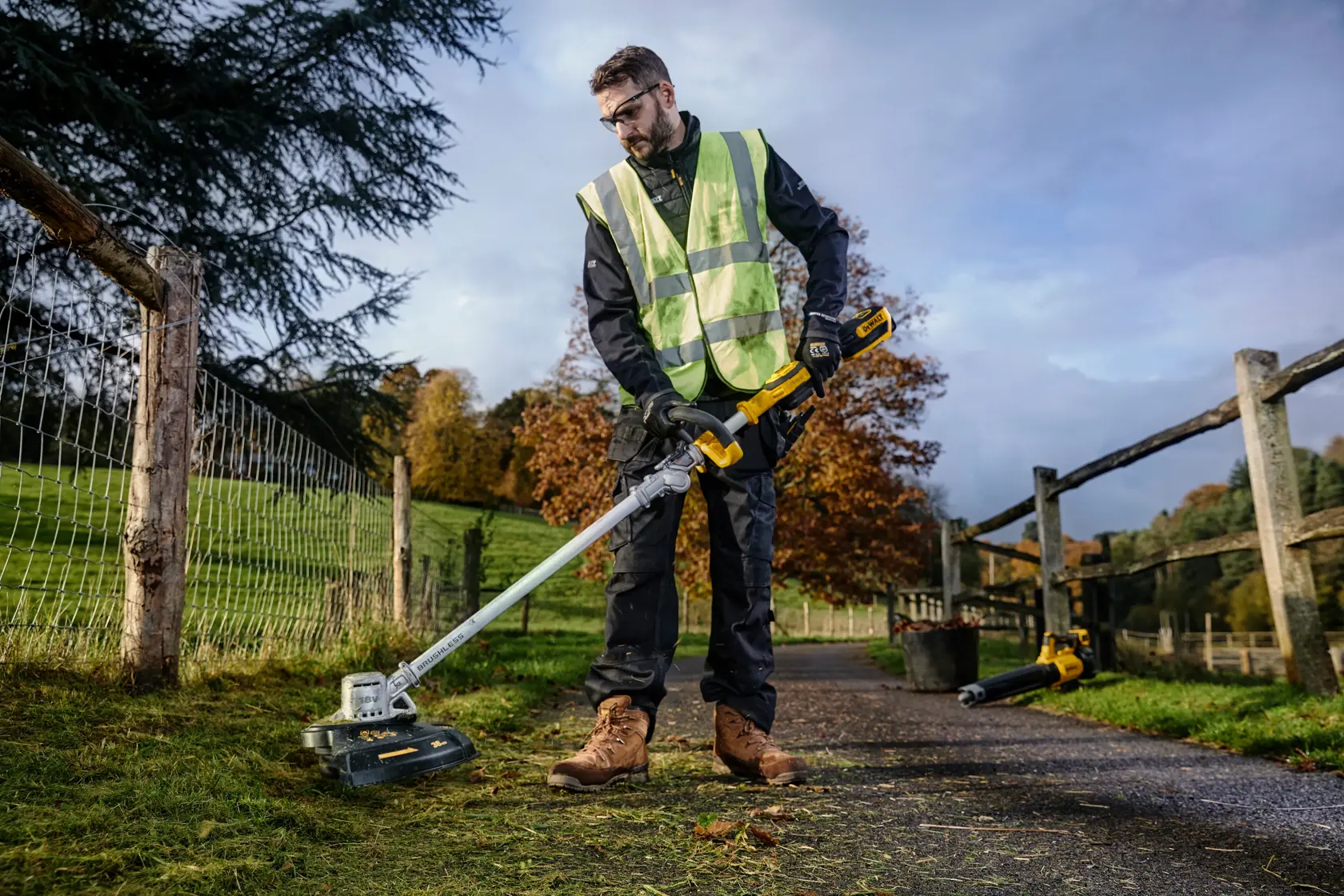 Person using a DEWALT cordless grass trimmer outdoors, wearing safety gear and working beside a wooden fence.