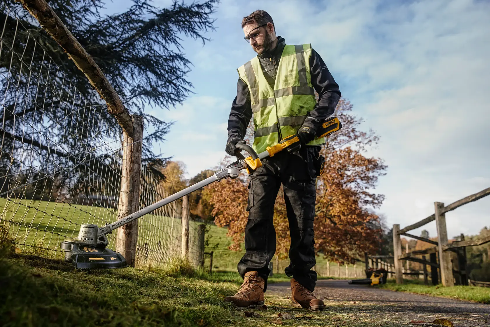 Person uses a DEWALT DCMST561P1 cordless string trimmer outdoors along a fence, wearing safety gear.