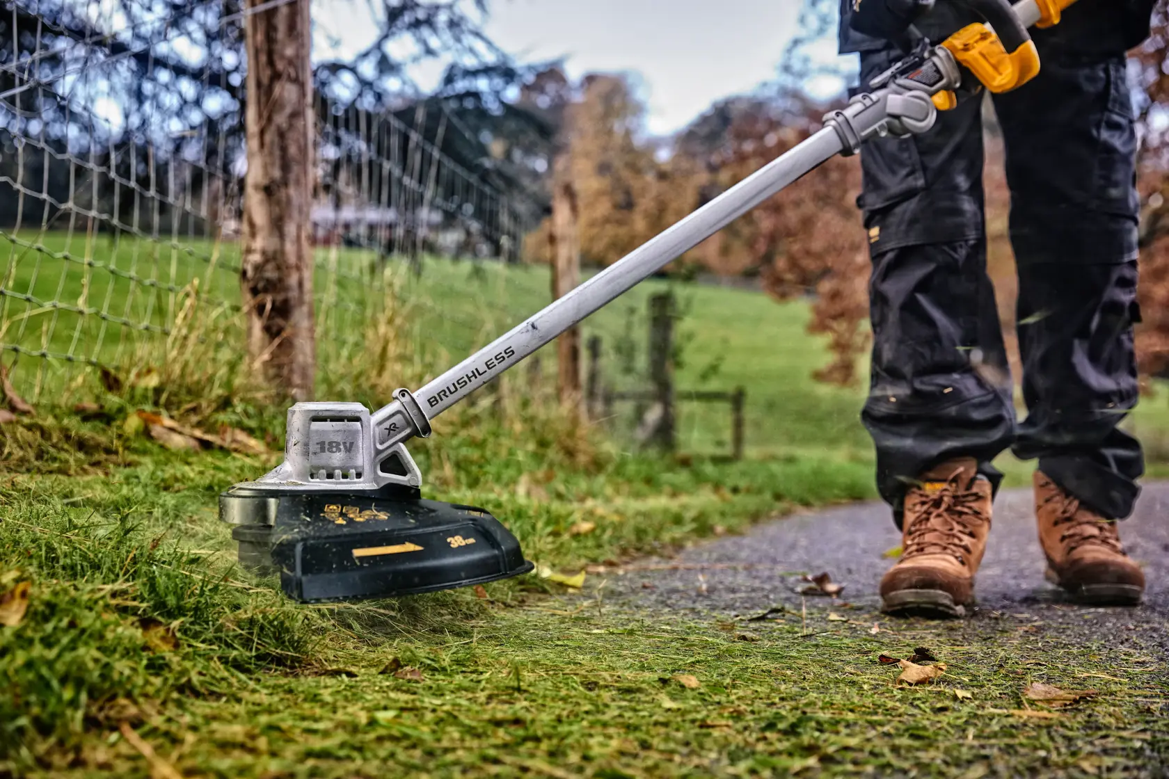 Person trims grass with a DEWALT cordless brushless string trimmer outdoors near a fence.