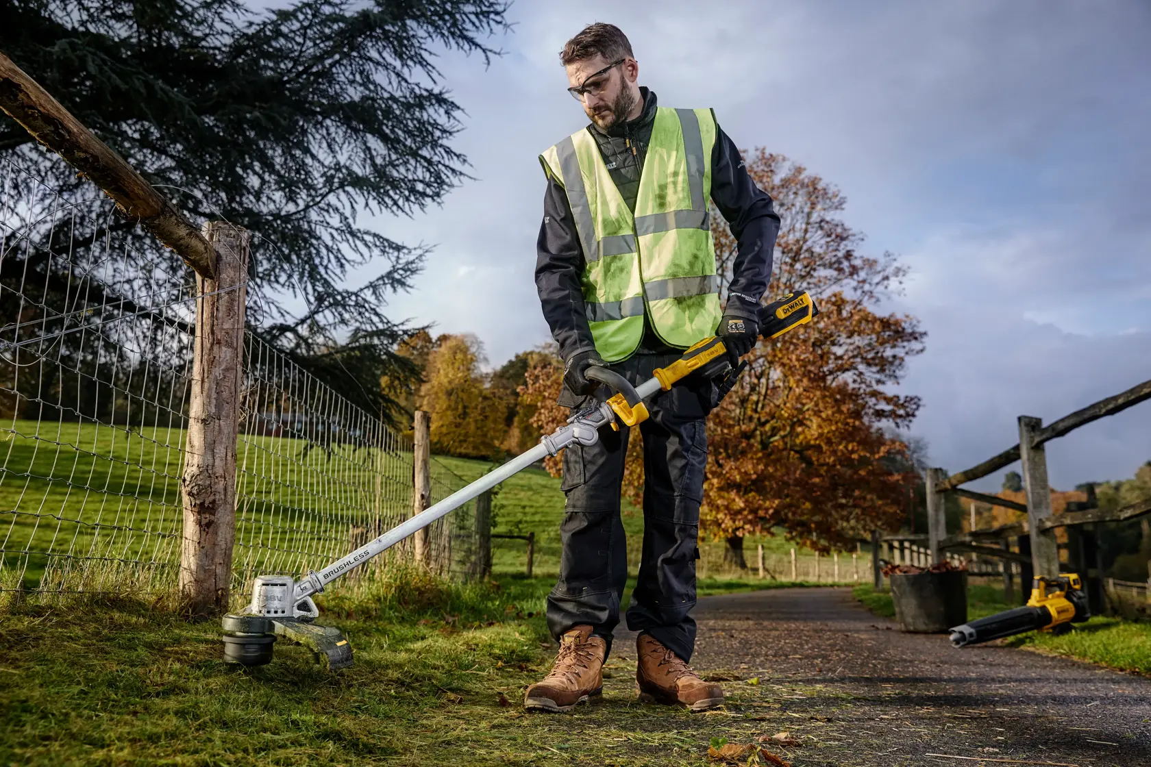 Person using a DEWALT cordless string trimmer outdoors near a fence in a grassy area.
