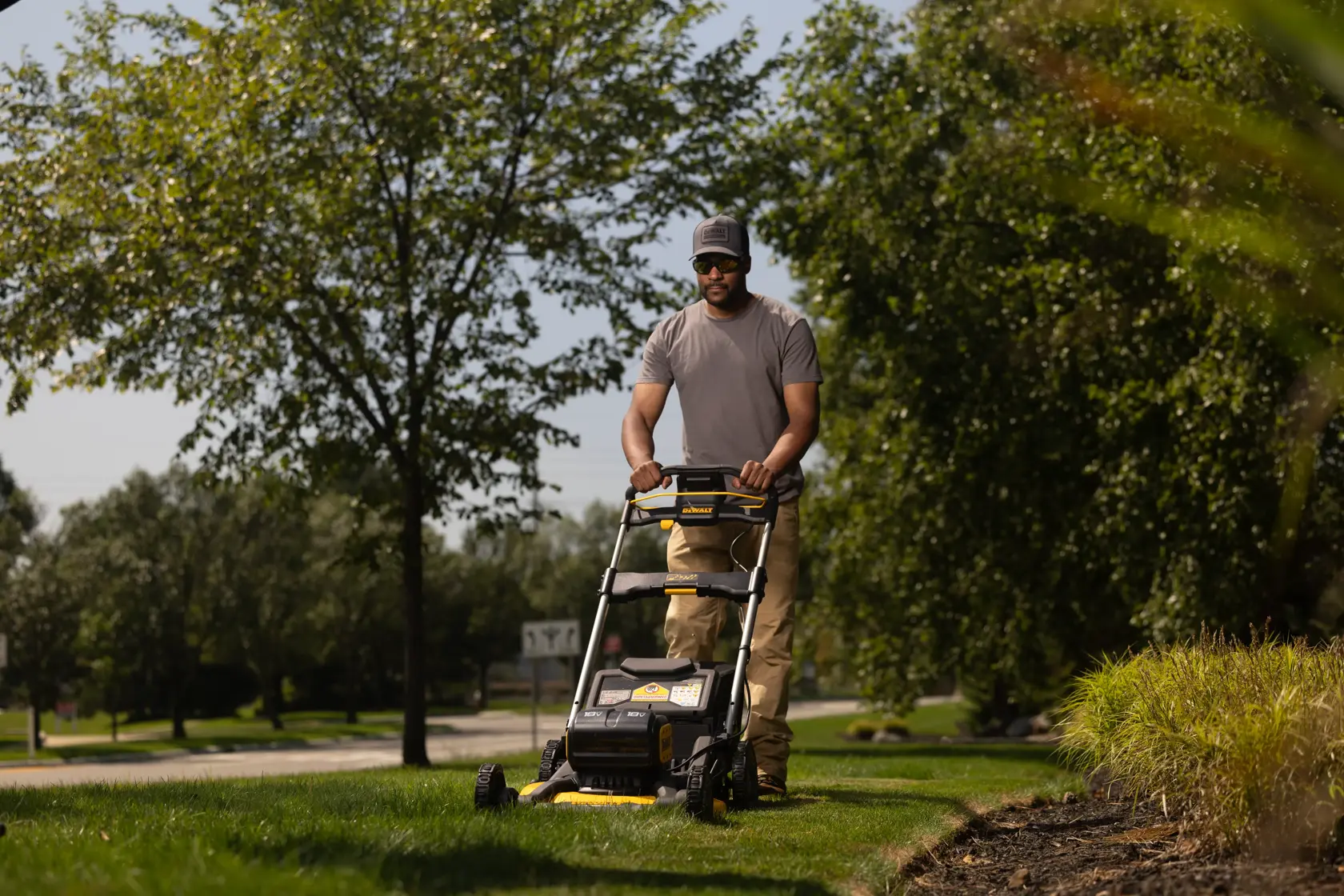 Una persona corta el césped con una cortadora de césped inalámbrica DEWALT modelo DCMWP134N, rodeado de árboles y plantas verdes en un día soleado.