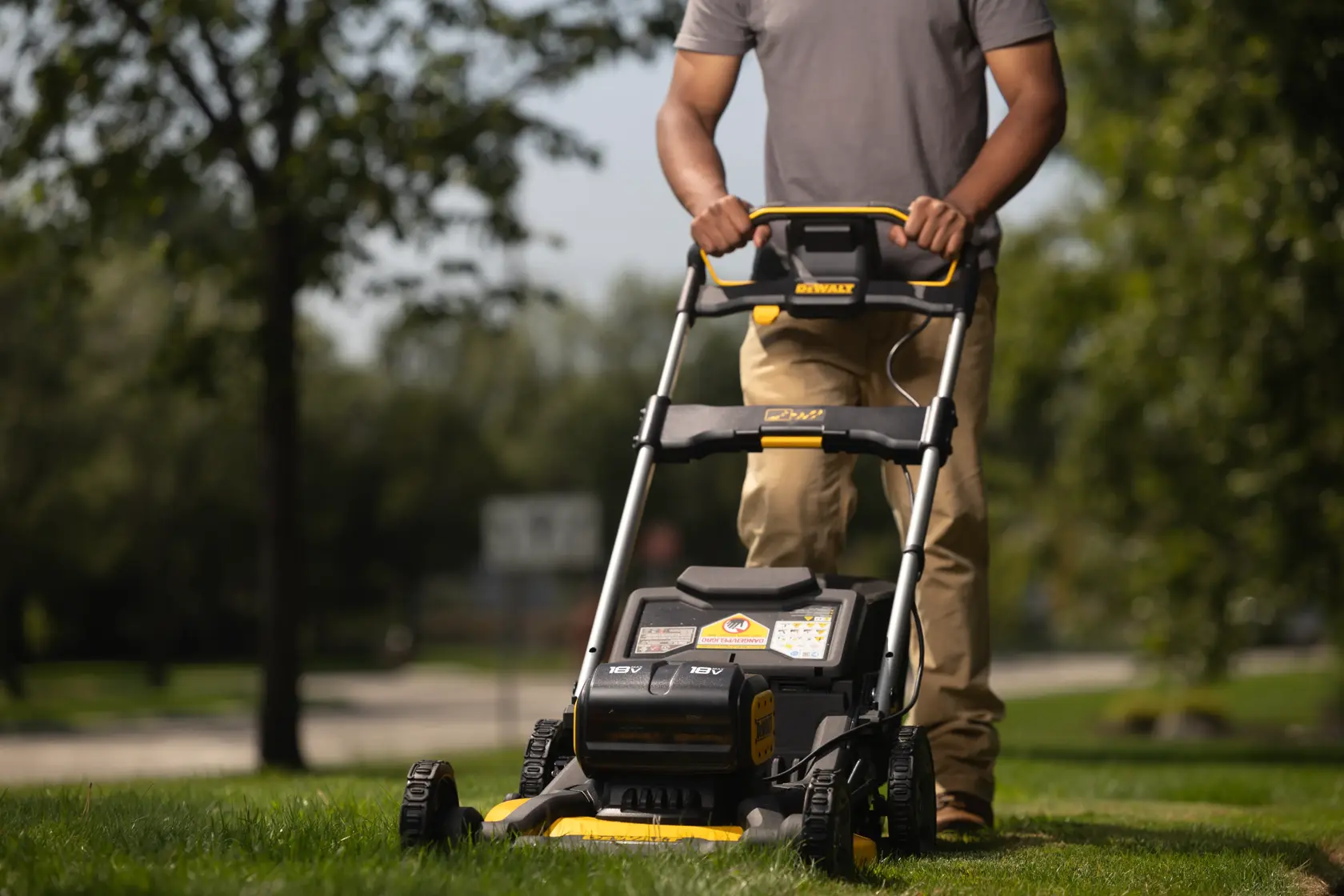 Una persona impulsa un cortacésped eléctrico inalámbrico DEWALT sobre césped verde al aire libre, con árboles y una carretera visible al fondo.