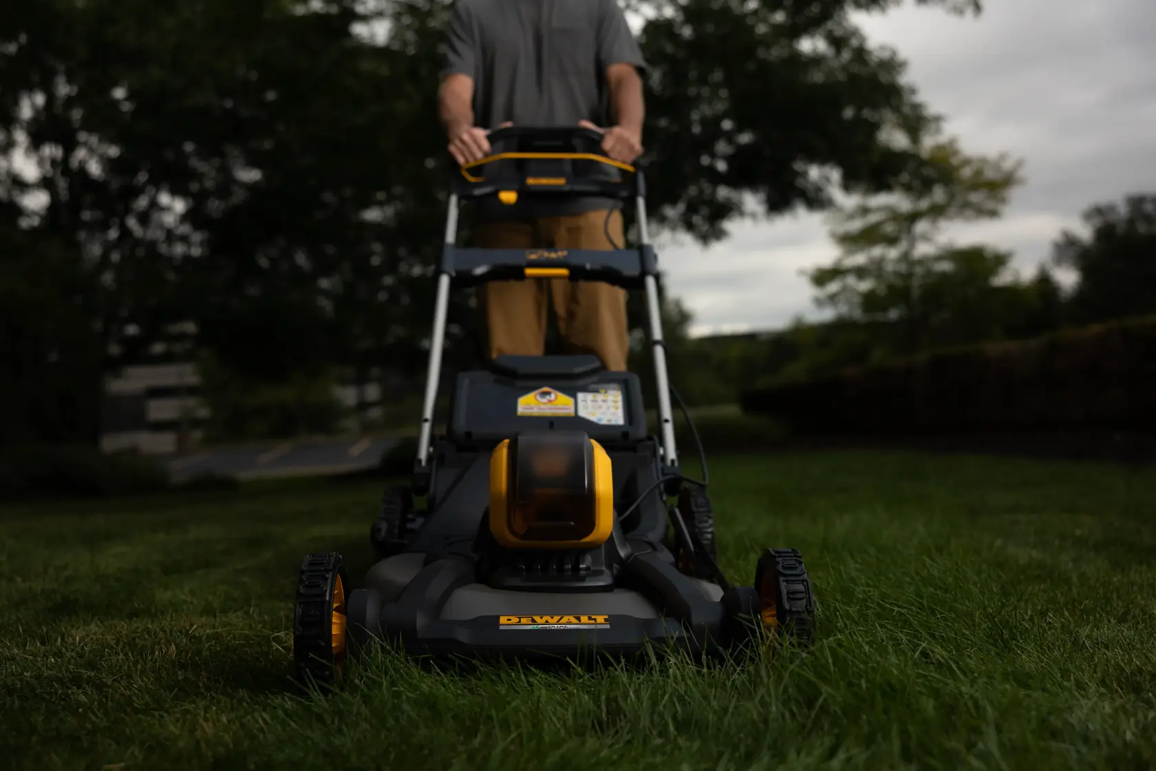 Ein kabelloser DEWALT-Rasenmäher wird im Freien über eine grasbewachsene Fläche geschoben, wobei eine Person dahinter steht