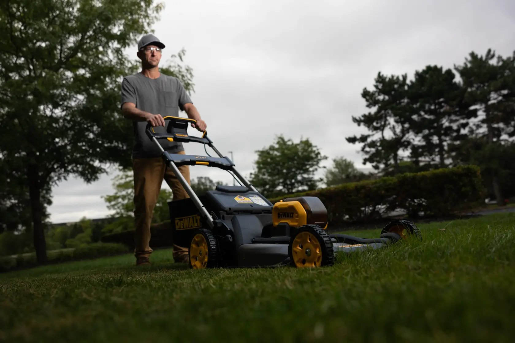 Eine Person nutzt einen DEWALT Akku-Rasenmäher im Freien, umgeben von Bäumen und Gras an einem wolkigen Tag.
