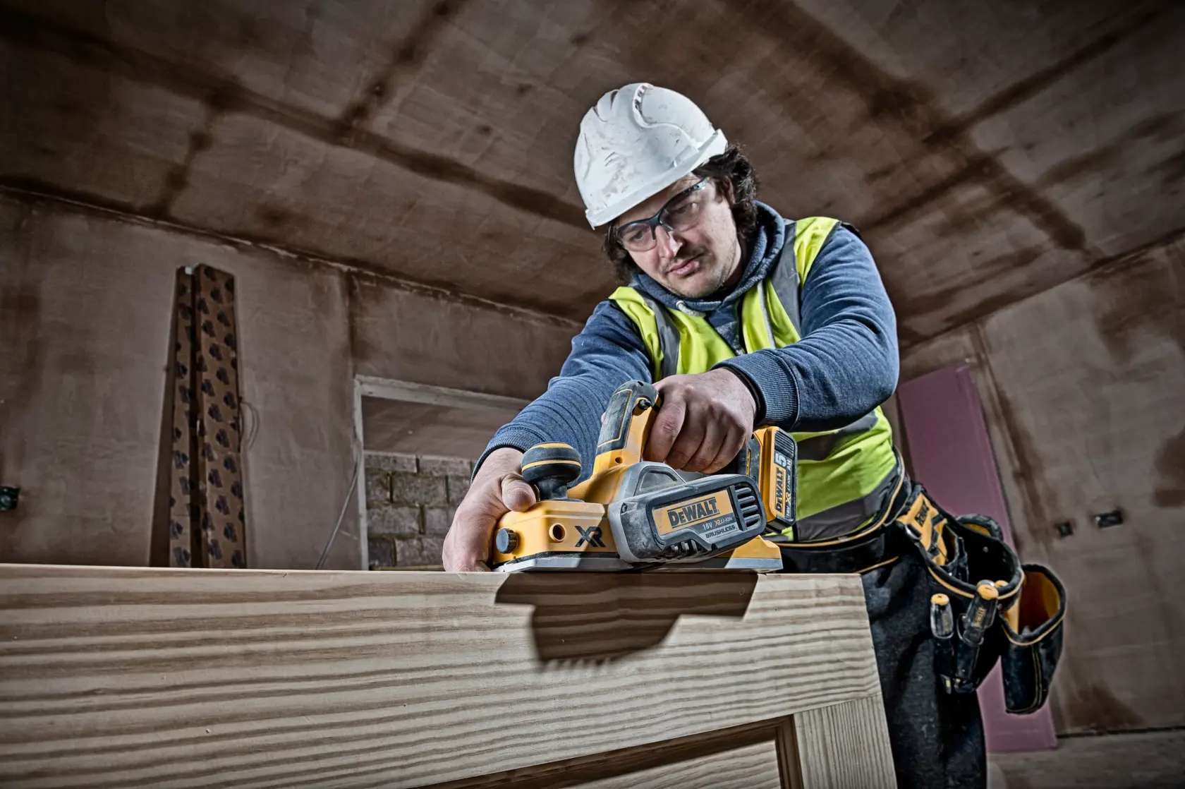 Worker uses DEWALT DCP580N planer on wood indoors, wearing safety gear and tool belt.