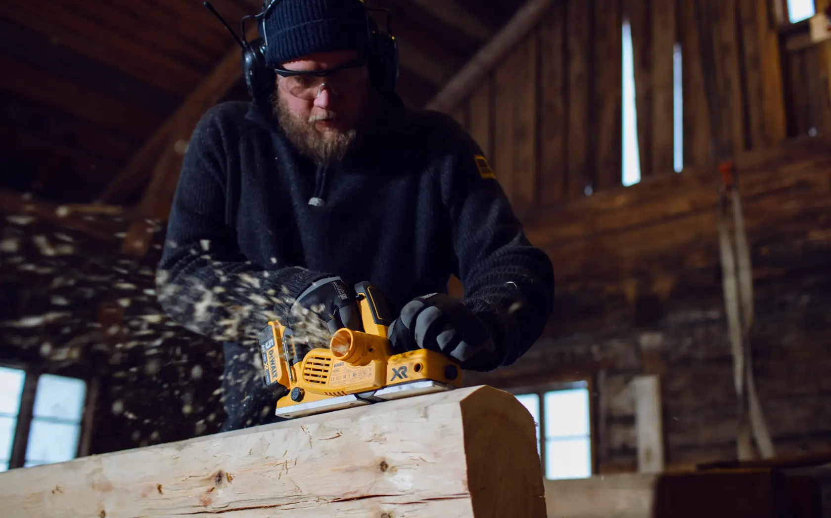 A person using a DEWALT DCP580P2 cordless planer to smooth a piece of wood inside a workshop, with wood shavings flying. The person's face is obscured and they are wearing protective headphones and gloves.