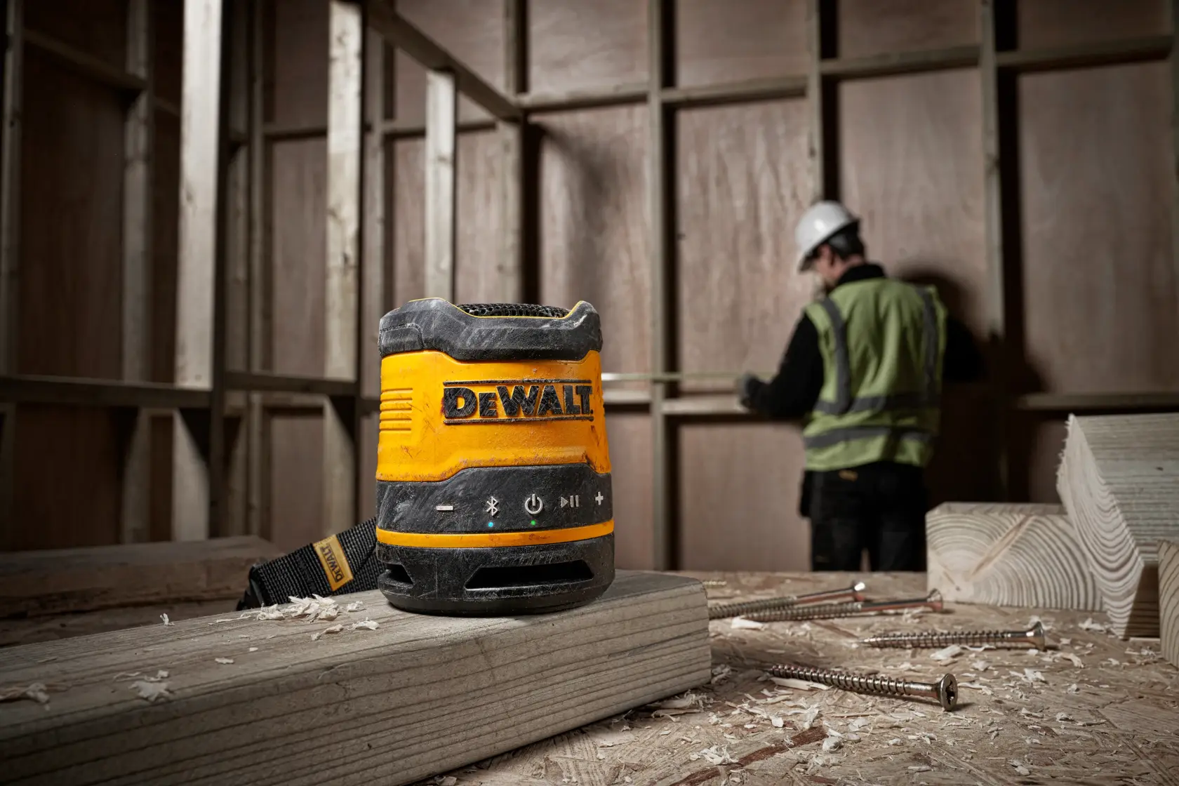 A DEWALT DCR009 portable Bluetooth speaker placed on a wooden plank in a construction site, with a worker in a safety vest and helmet measuring in the background.