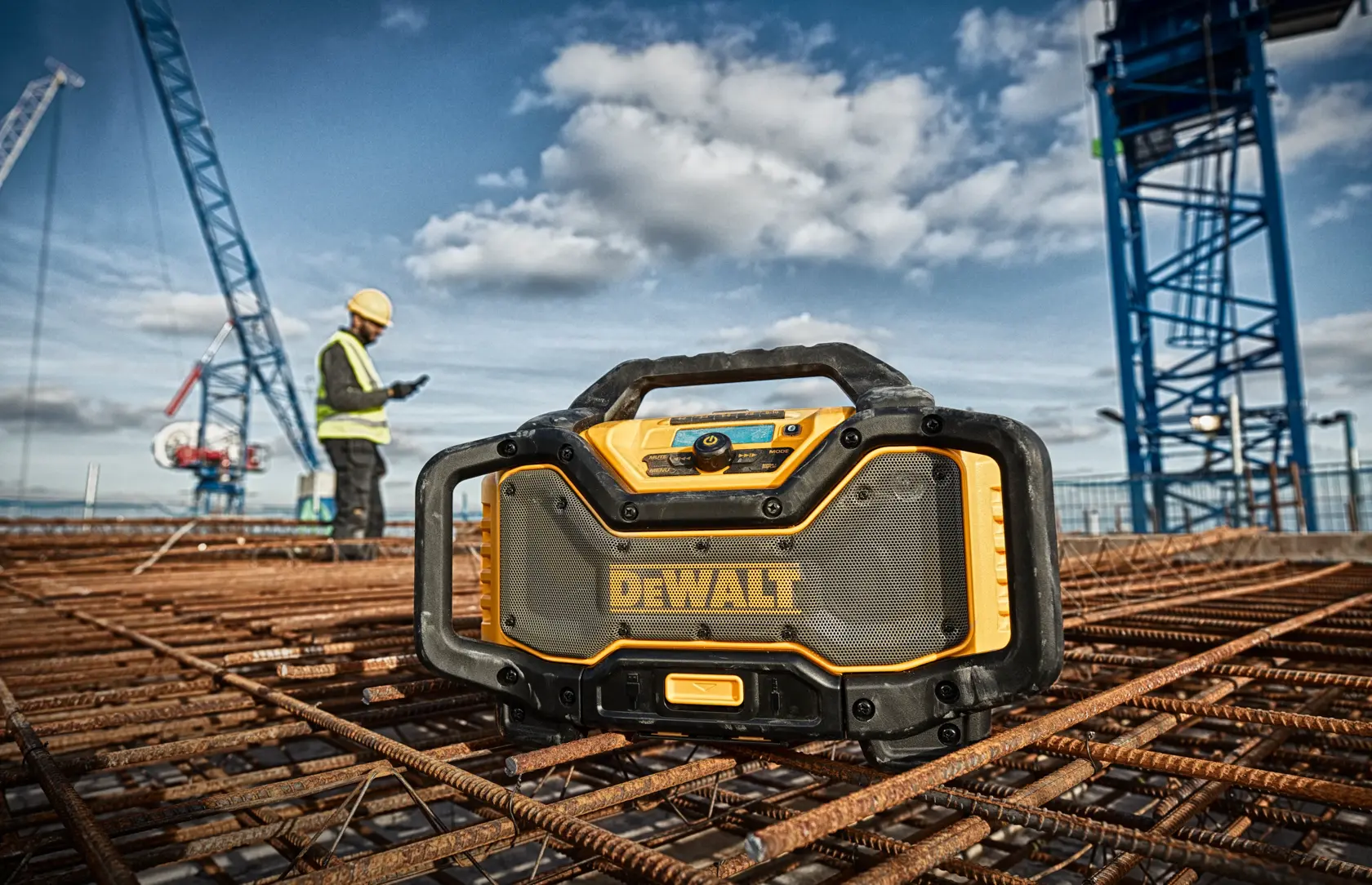 DEWALT Baustellenradio auf Bewehrung, mit Arbeiter und Kränen im Hintergrund auf einer Baustelle.