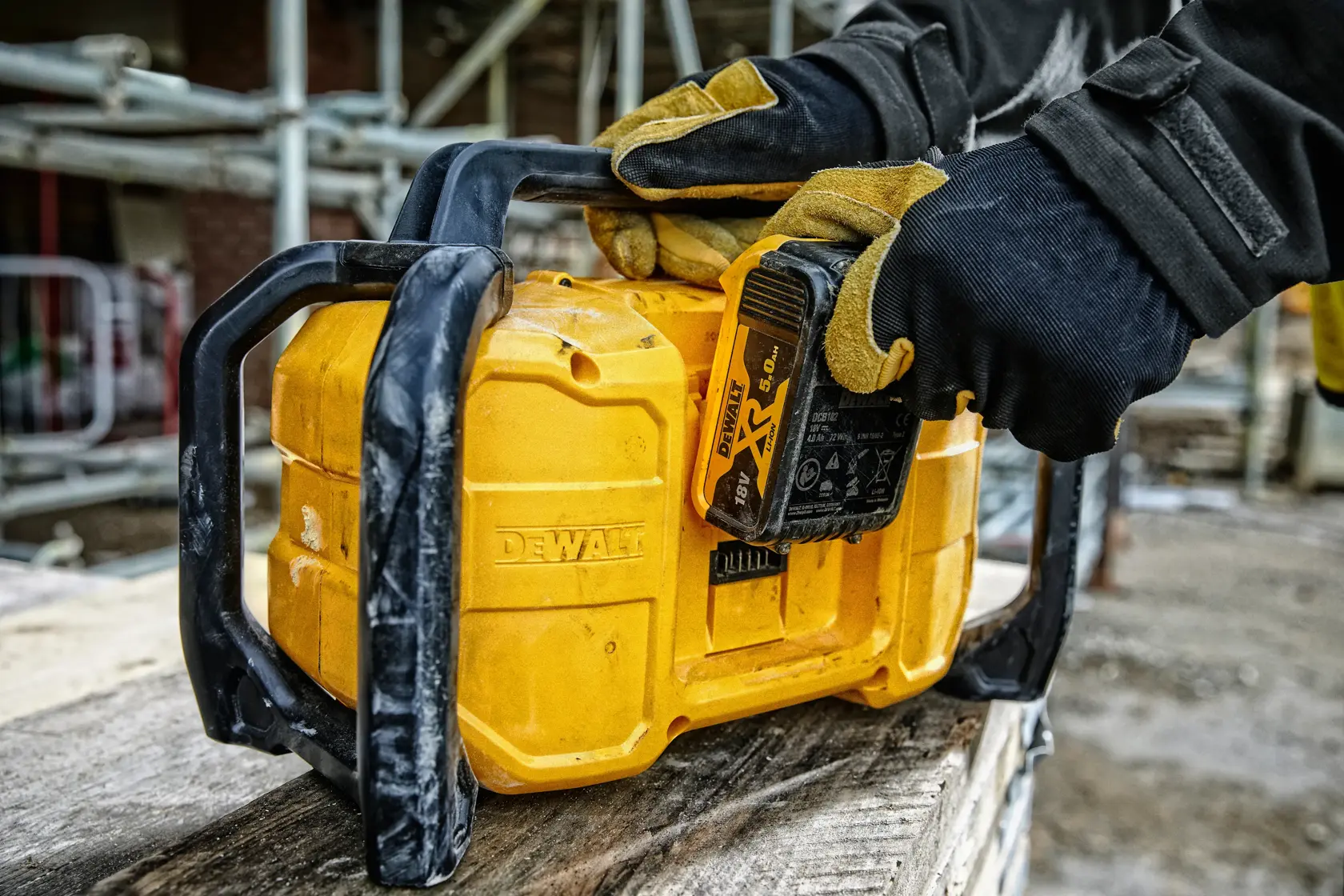 Handschuhhände setzen Batterie in gelbes DEWALT-Gerät auf Baustelle mit Gerüst im Hintergrund ein.