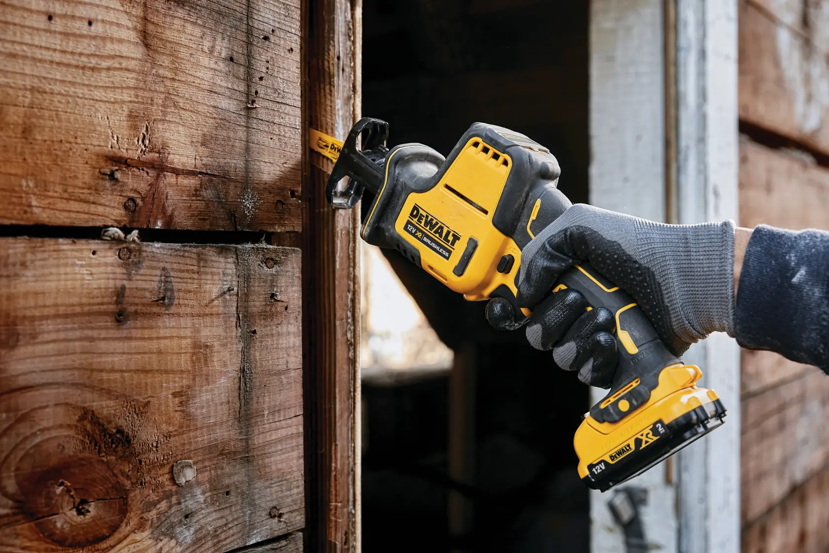 Sierra recíproca inalámbrica DEWALT cortando madera, sostenida por una mano enguantada junto a pared de madera.