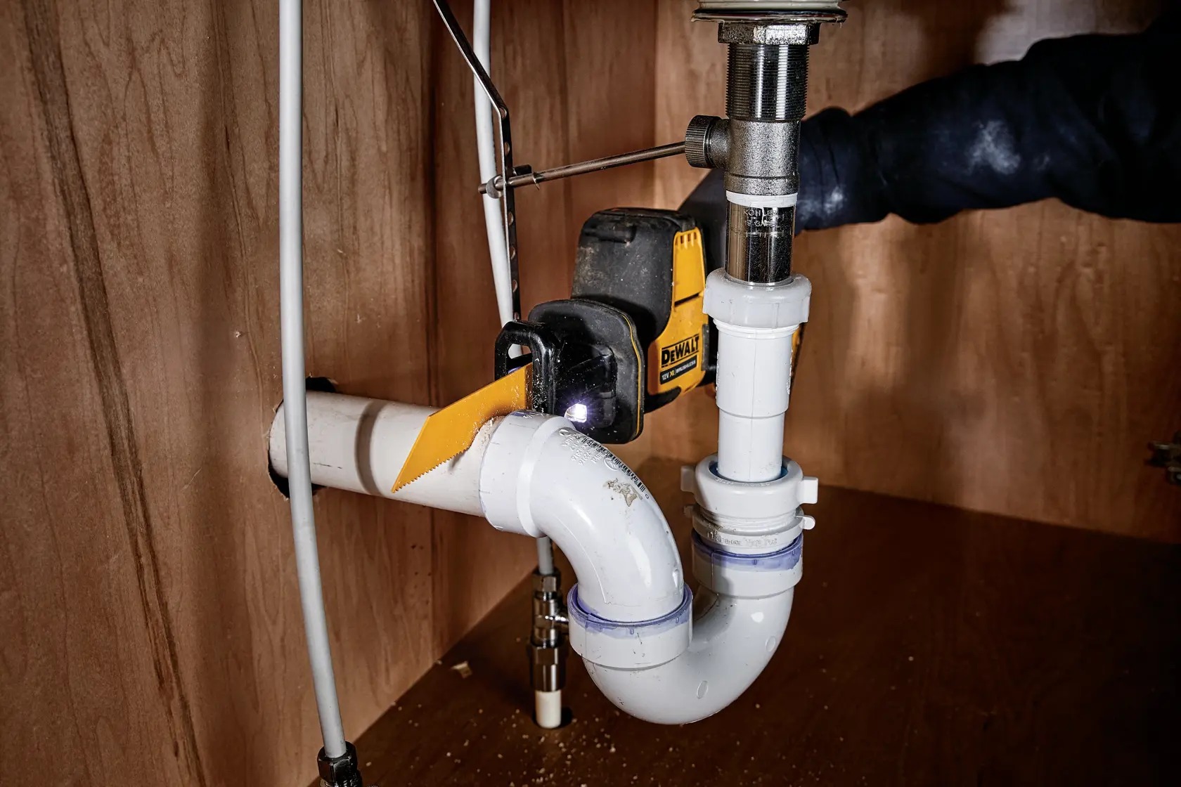 Sierra inalámbrica DEWALT cortando tubo de PVC bajo lavabo en gabinete de madera.