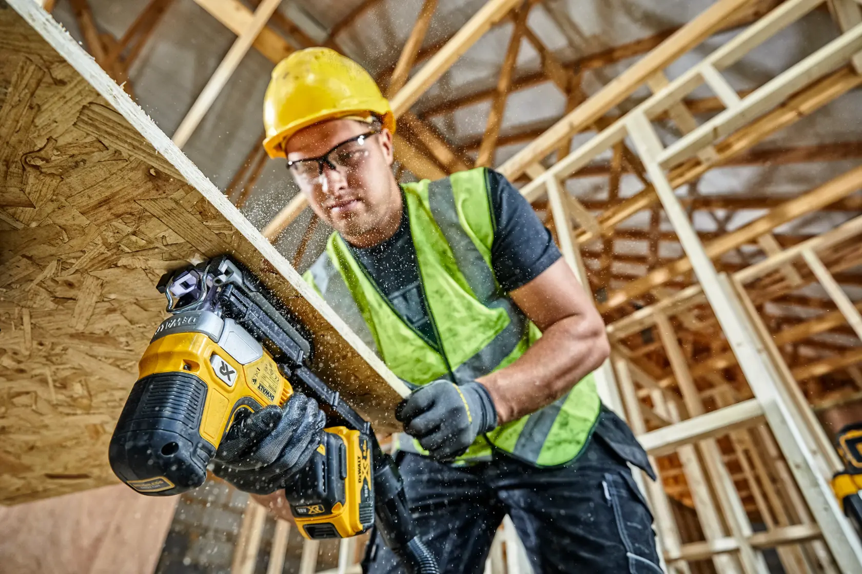 Worker uses DEWALT DCS335P2 jigsaw to cut wood at a construction site, wearing safety gear and a yellow hard hat.