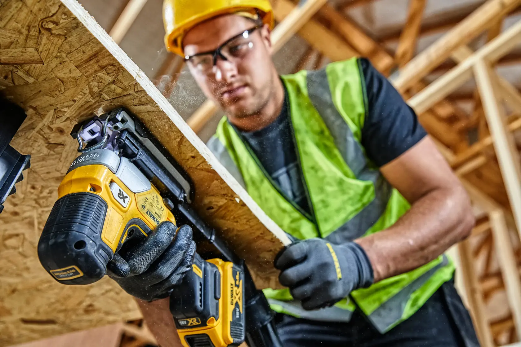 Worker uses a DEWALT cordless jigsaw to cut wood panel at a construction site, wearing safety gear.