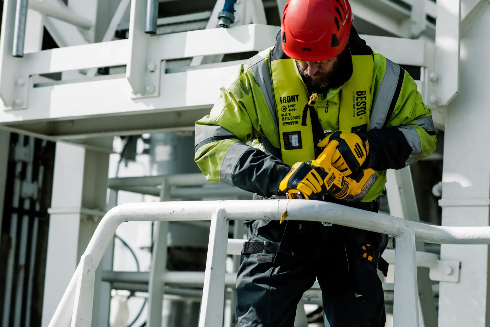 A person wearing a red safety helmet and a high-visibility jacket operates a yellow DEWALT power tool to cut metal railings at an industrial worksite.