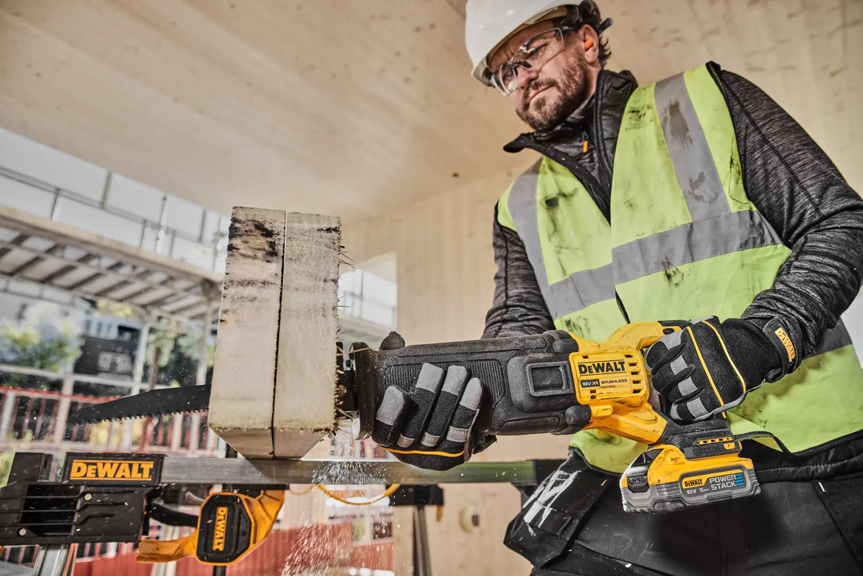 A construction worker wearing safety gear, including a helmet, gloves, and a high-visibility vest, is using a DEWALT cordless reciprocating saw (model DCS386H2T) to cut through a thick piece of wood on a building site.