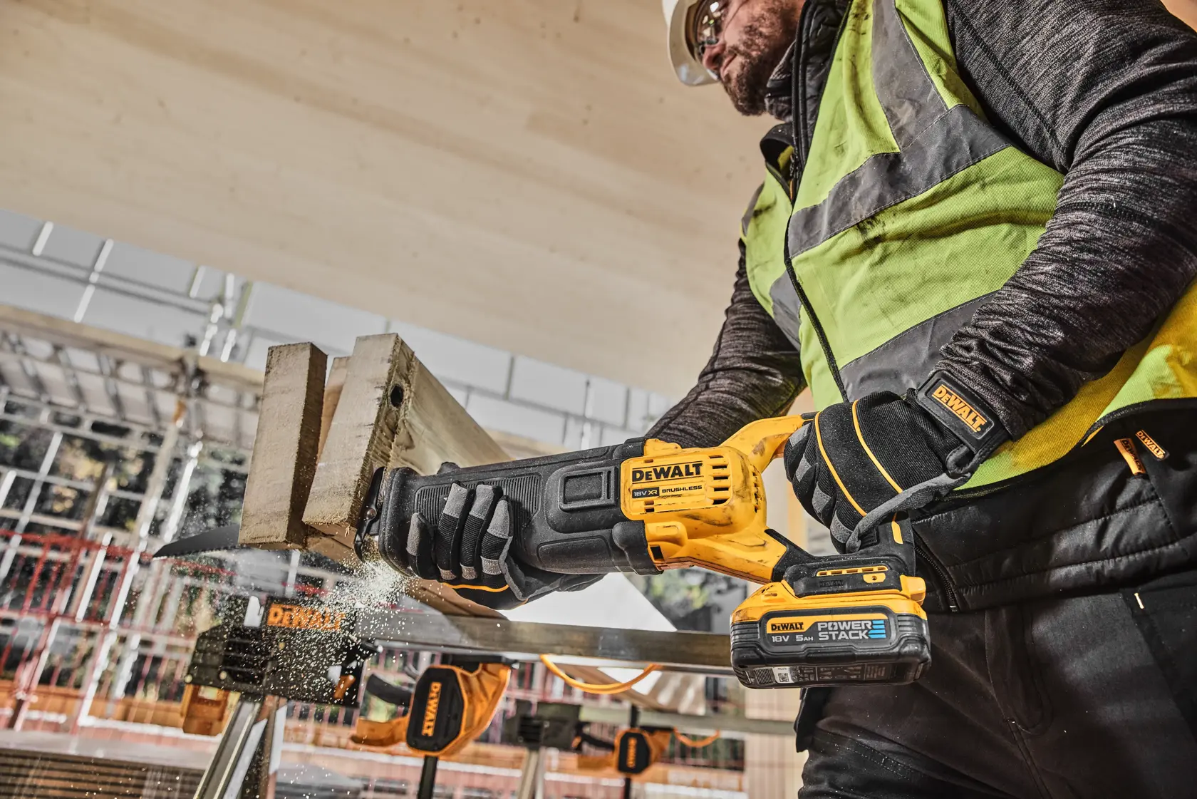 A construction worker using a DEWALT DCS386H2T cordless reciprocating saw to cut a piece of wood at a building site, wearing safety gear including gloves, vest, and helmet.