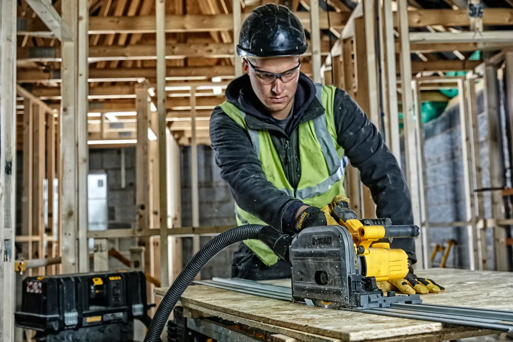 Arbeiter schneidet Holz mit DEWALT DCS520NT Tauchsäge auf Baustelle. Schutzkleidung und Werkzeuge sichtbar.