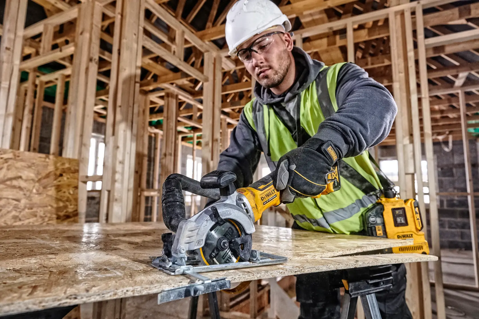 Person uses DEWALT DCS571N saw to cut wood sheet at construction site, wearing safety gear and high-vis vest.