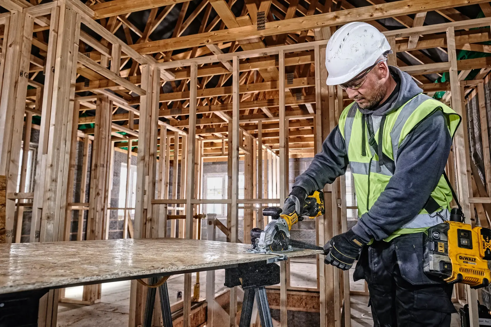 Worker uses DEWALT DCS571N saw to cut wood panel at construction site, wearing safety gear and high-visibility vest.