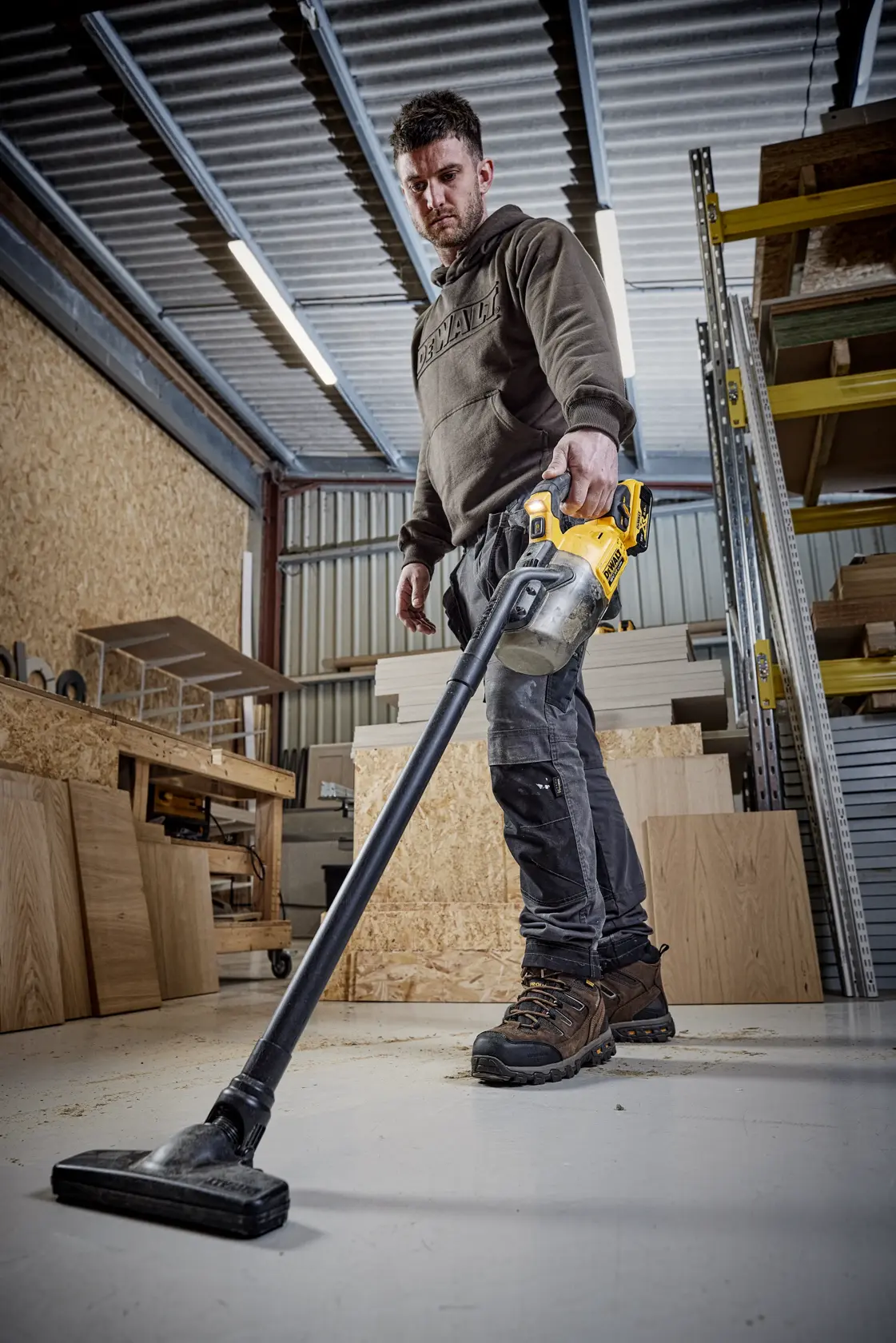 Persona usando aspiradora DEWALT DCV501LN en el taller, entre paneles de madera y estanterías.