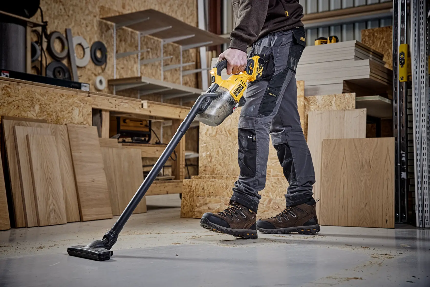 Persona usando aspiradora inalámbrica DEWALT para limpiar aserrín en el suelo de taller, rodeada de paneles de madera.