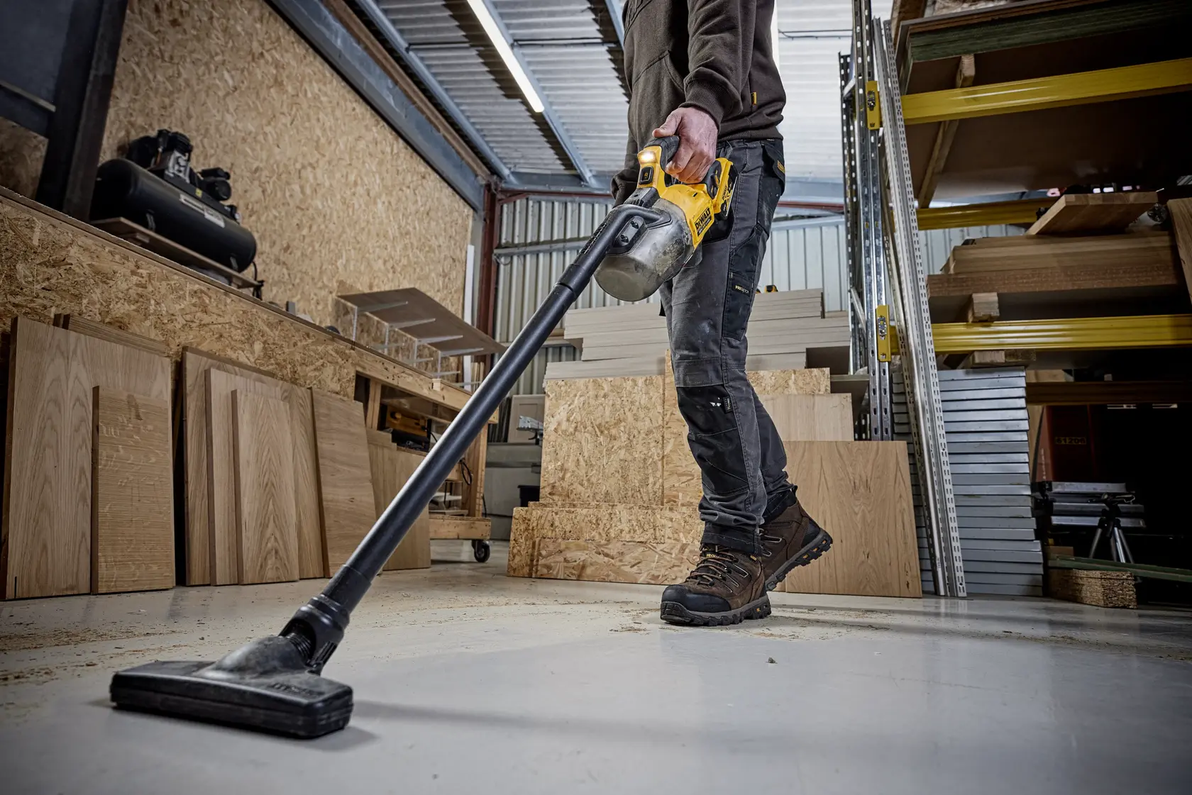 Persona limpia el suelo del taller con aspiradora inalámbrica DEWALT; hay tablas y estantes visibles.