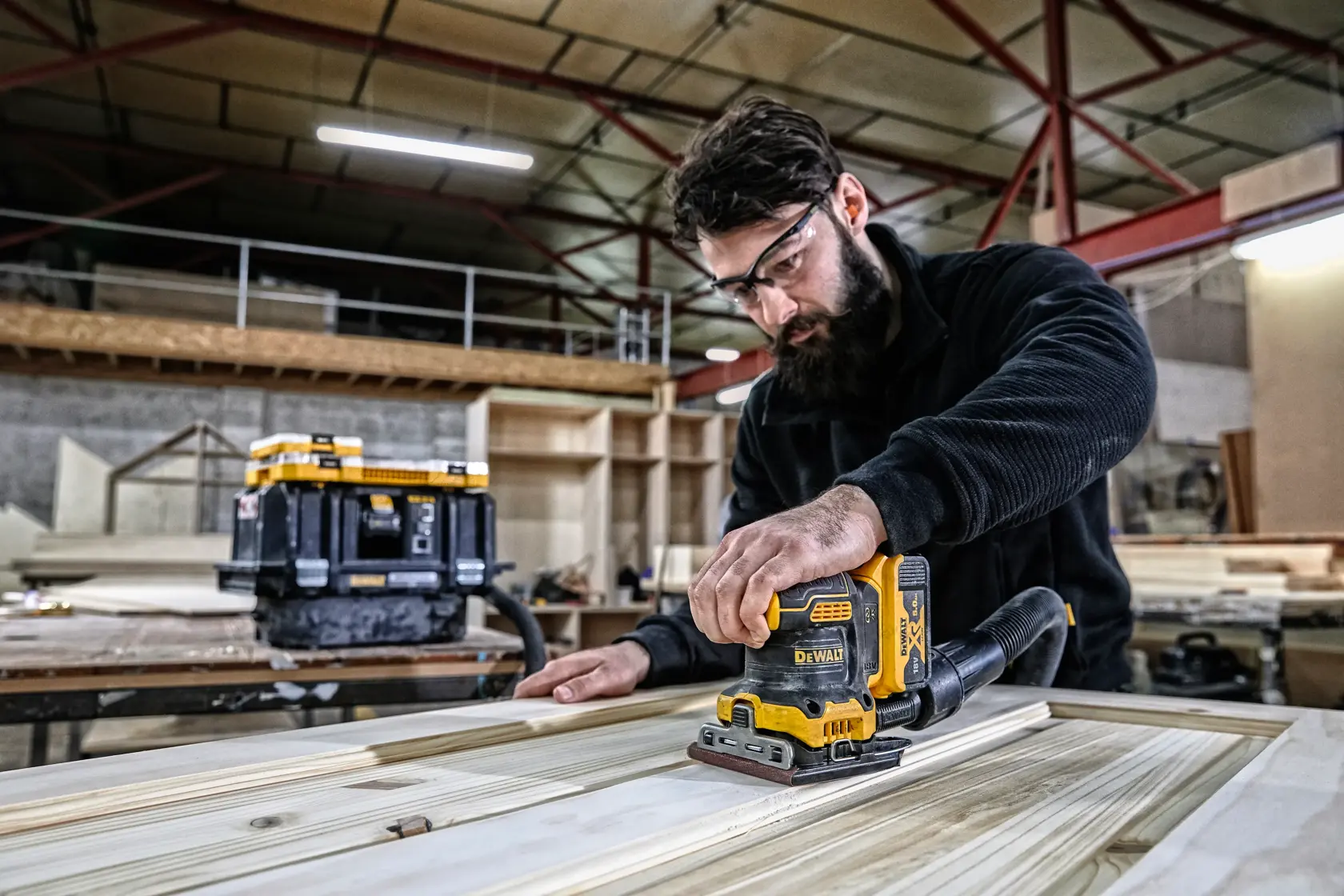 Persona lijando madera con lijadora DEWALT DCW200P2 en taller. Caja de herramientas al fondo.