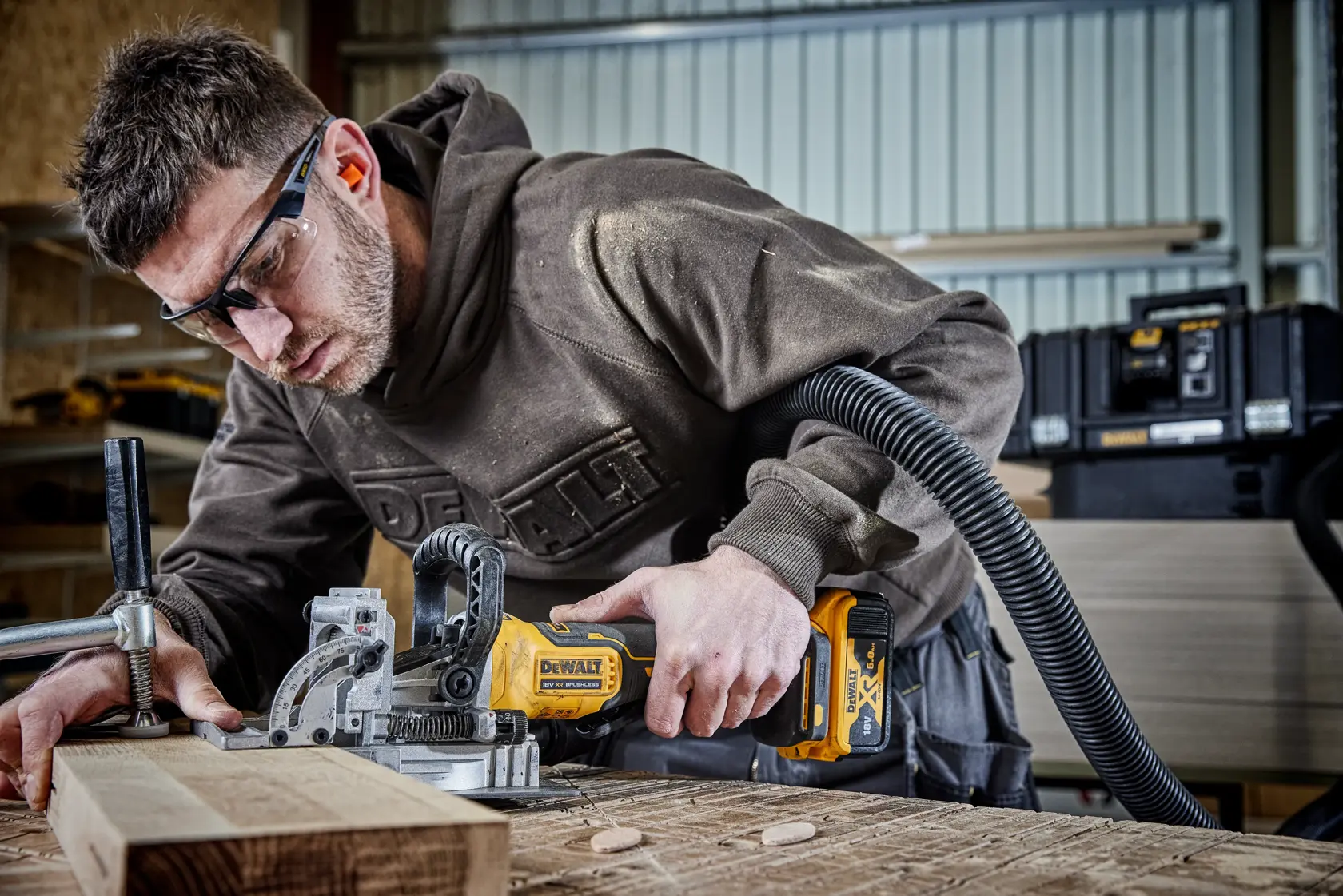 Person uses DEWALT cordless joiner to cut wood in a workshop, with dust extractor attached and toolboxes in background.