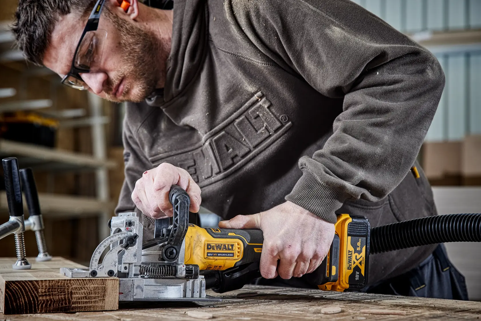 Person using a DEWALT DCW682N cordless jointer on wood in a workshop, attached to a dust extraction hose.