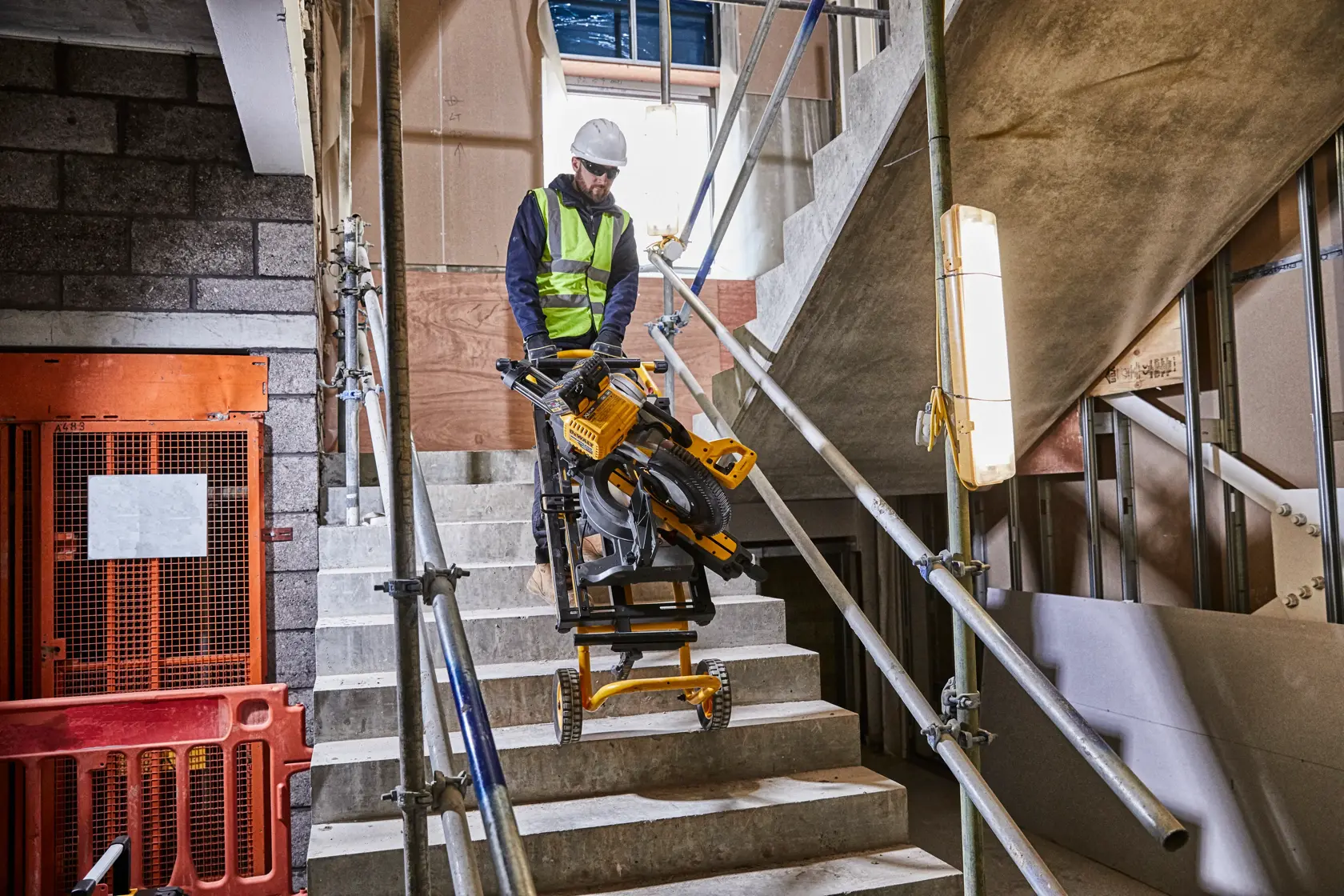 Trabajador mueve sierra y soporte DEWALT por escaleras de obra con barandillas y barreras visibles.