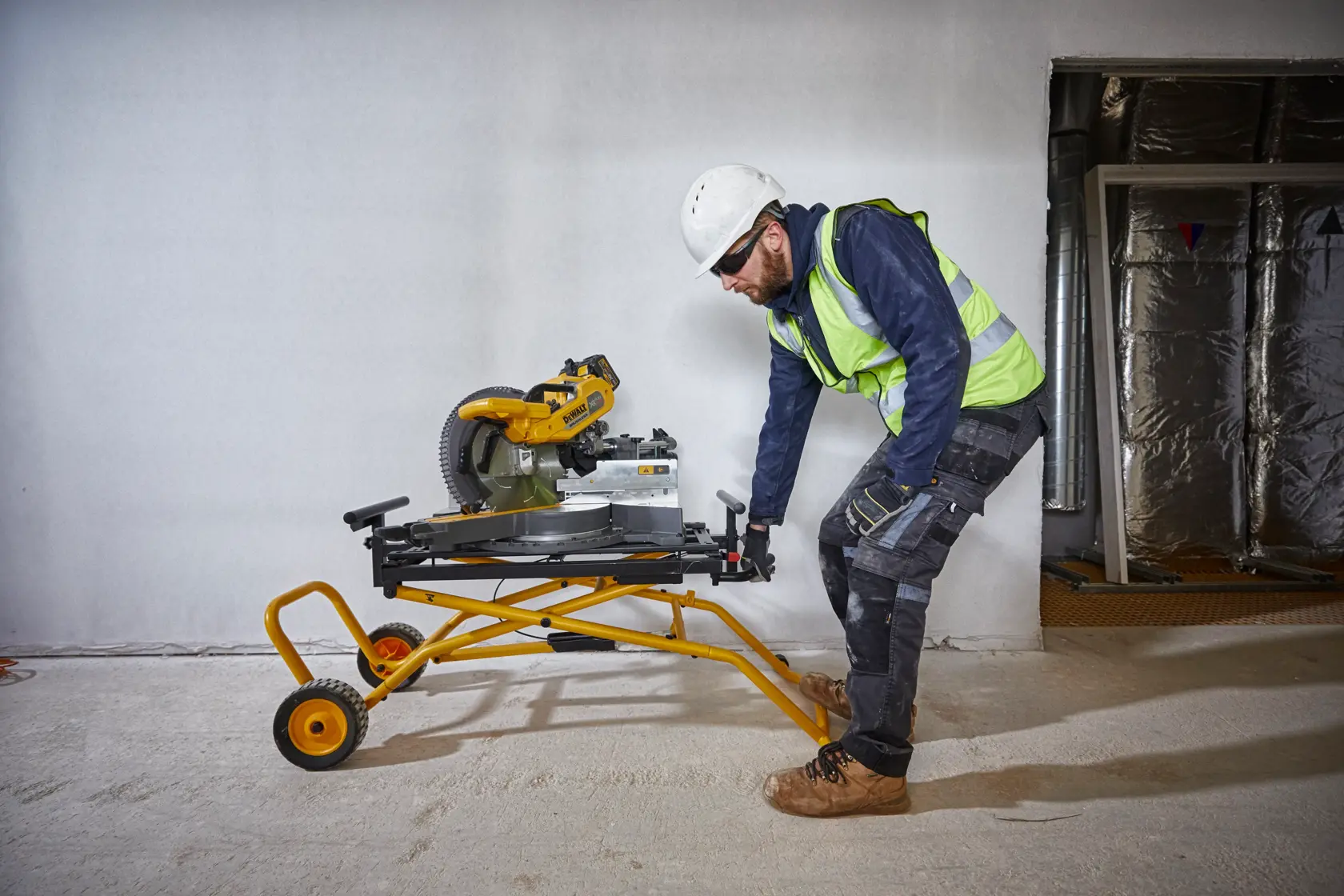 Trabajador mueve una sierra DEWALT sobre soporte amarillo con ruedas en obra.