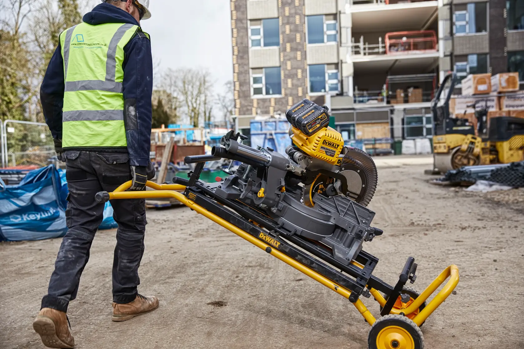 Trabajador mueve una ingletadora DEWALT en soporte con ruedas en obra, edificios al fondo.