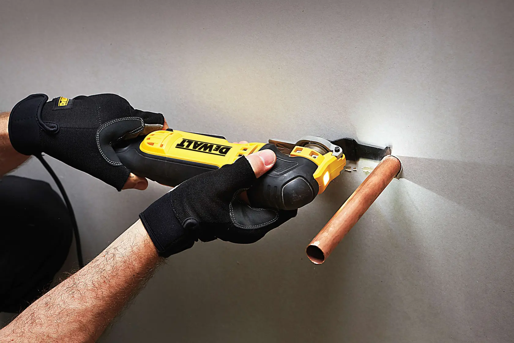 Person schneidet mit DEWALT Werkzeug eine Wand nahe Kupferrohr und trägt schwarze Handschuhe.
