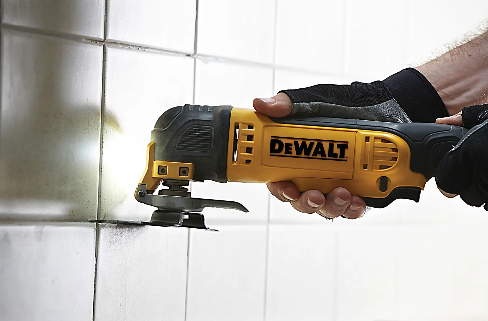 DEWALT oscillating multi-tool cutting tile grout, held by gloved hand against white tiled wall.