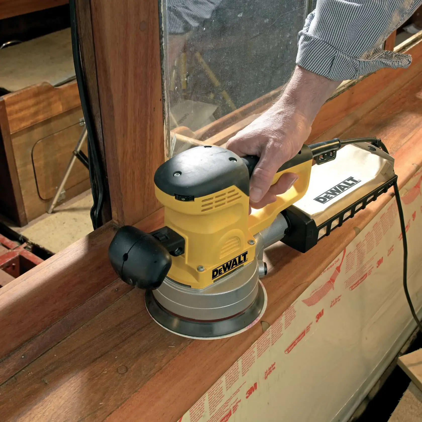 Person uses a DEWALT DT3123 sander to smooth wooden surface near a window. Tool is powered and yellow-black.