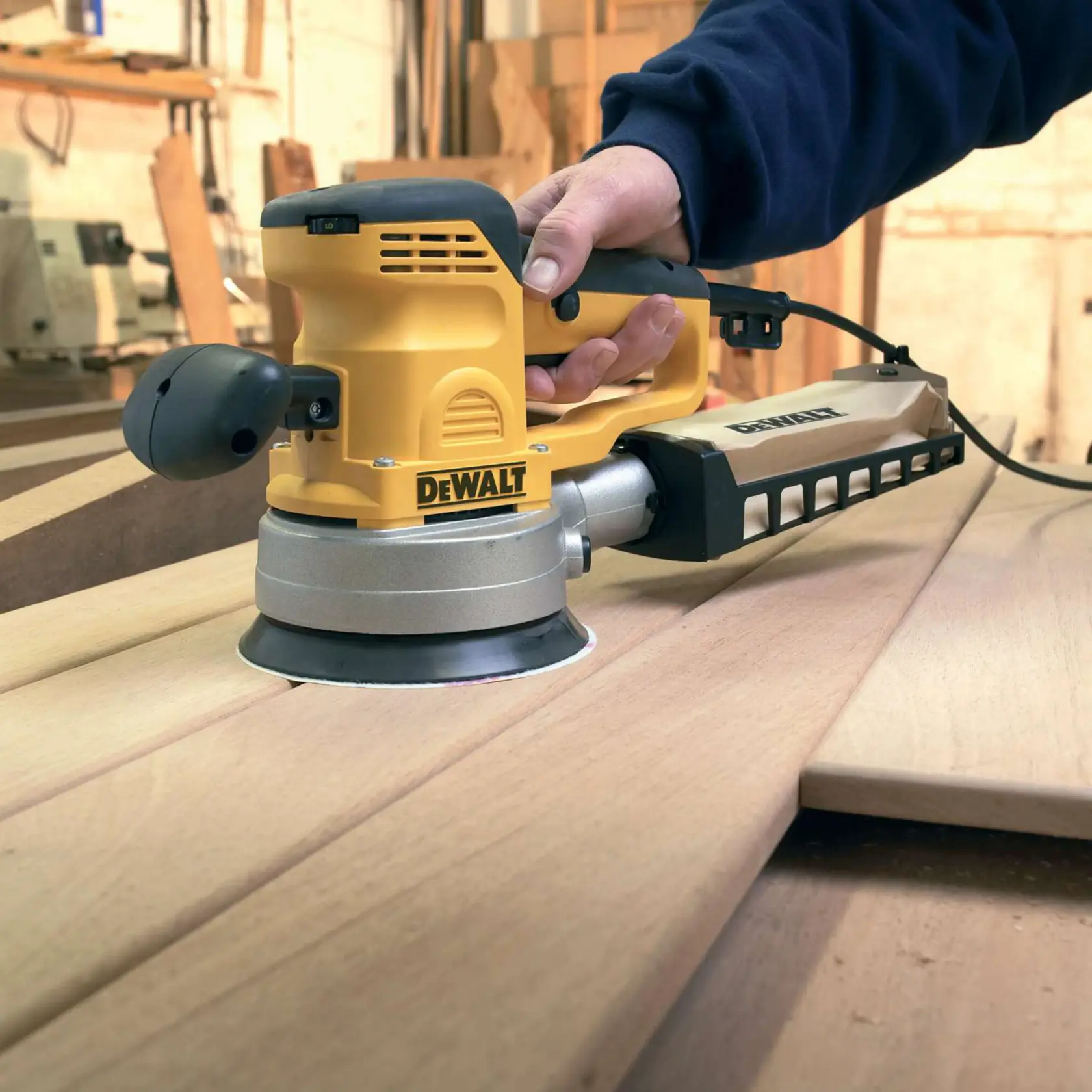 DEWALT orbital sander in use on wooden boards in a workshop, held by a person wearing a dark sleeve.