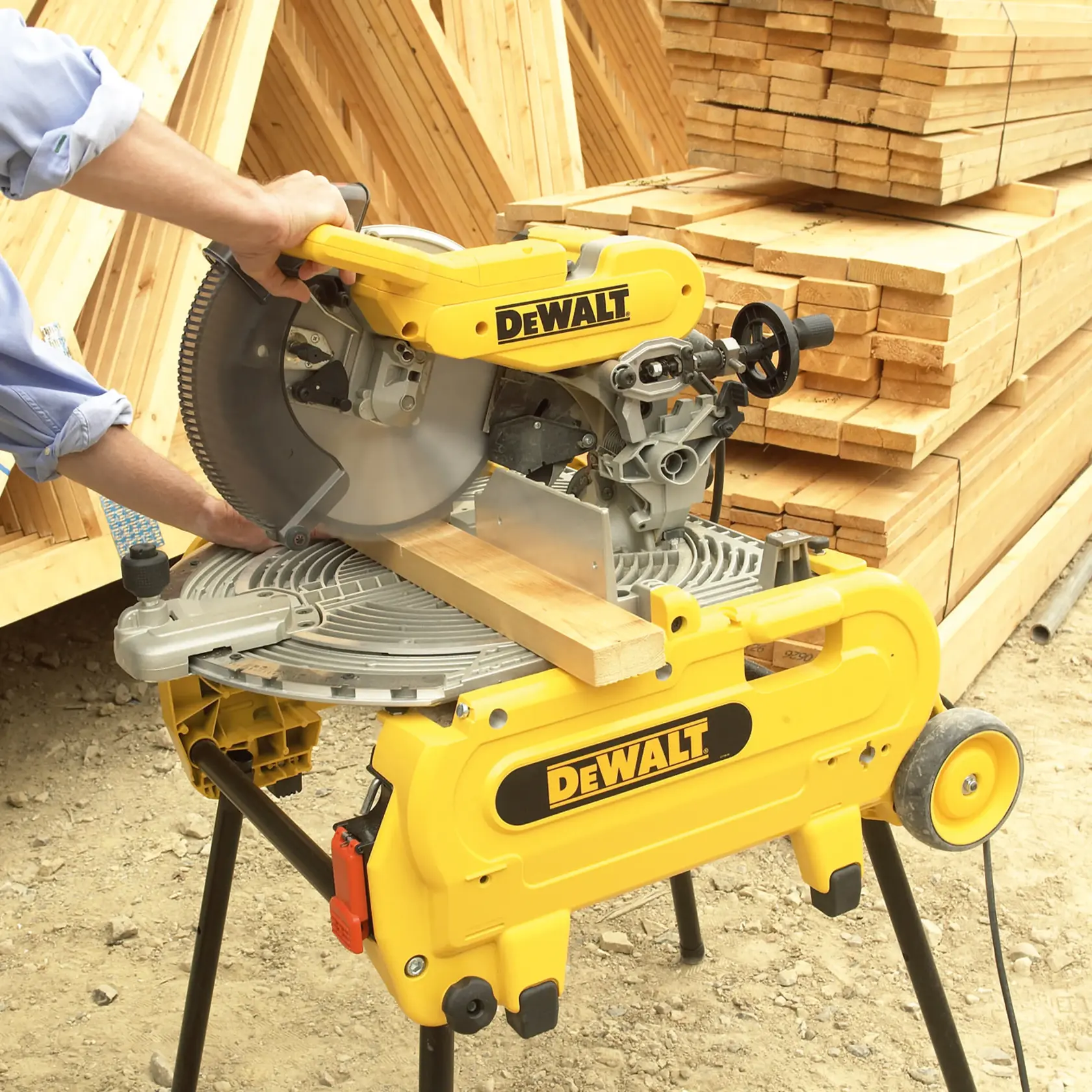 Una sierra de inglete DEWALT se está usando para cortar una pieza de madera, colocada sobre un soporte amarillo DEWALT. Se ven pilas de tablones de madera en el fondo de una obra exterior.