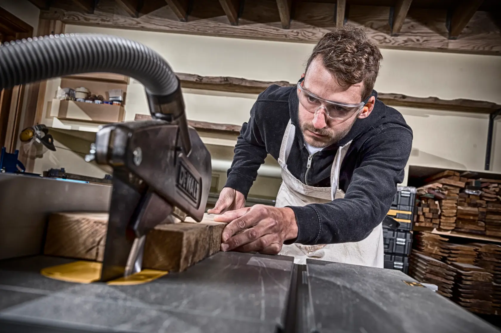Persona corta madera con una sierra DEWALT en taller, viste delantal. Pilas de madera en el fondo.