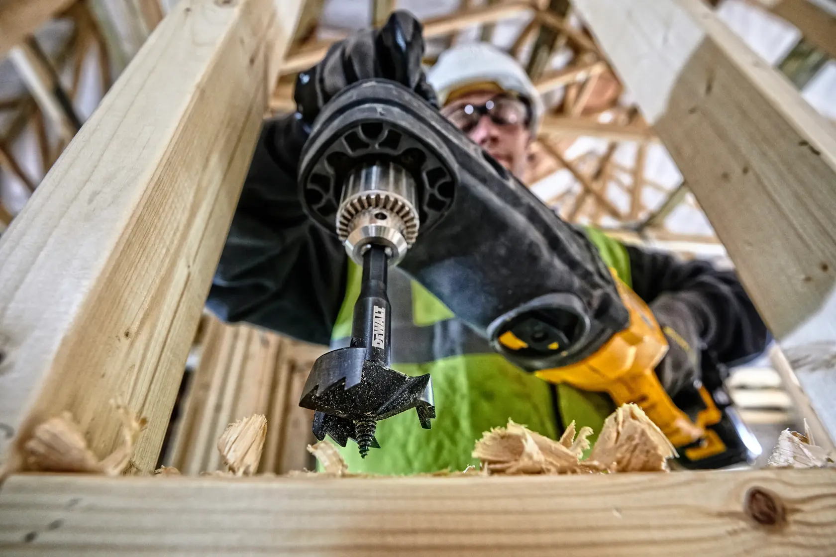 Primer plano de taladro DEWALT haciendo agujero en madera, virutas visibles, persona con chaleco verde.