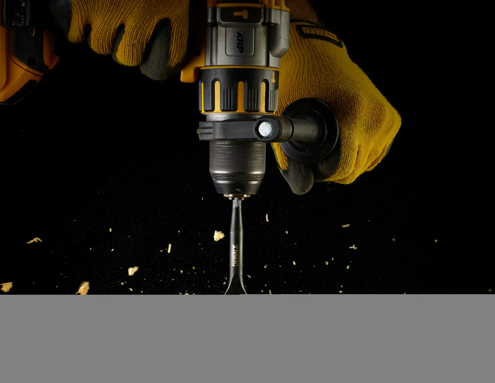 Gloved hands using a DEWALT drill and spade bit to drill into wood with scattered wood chips.